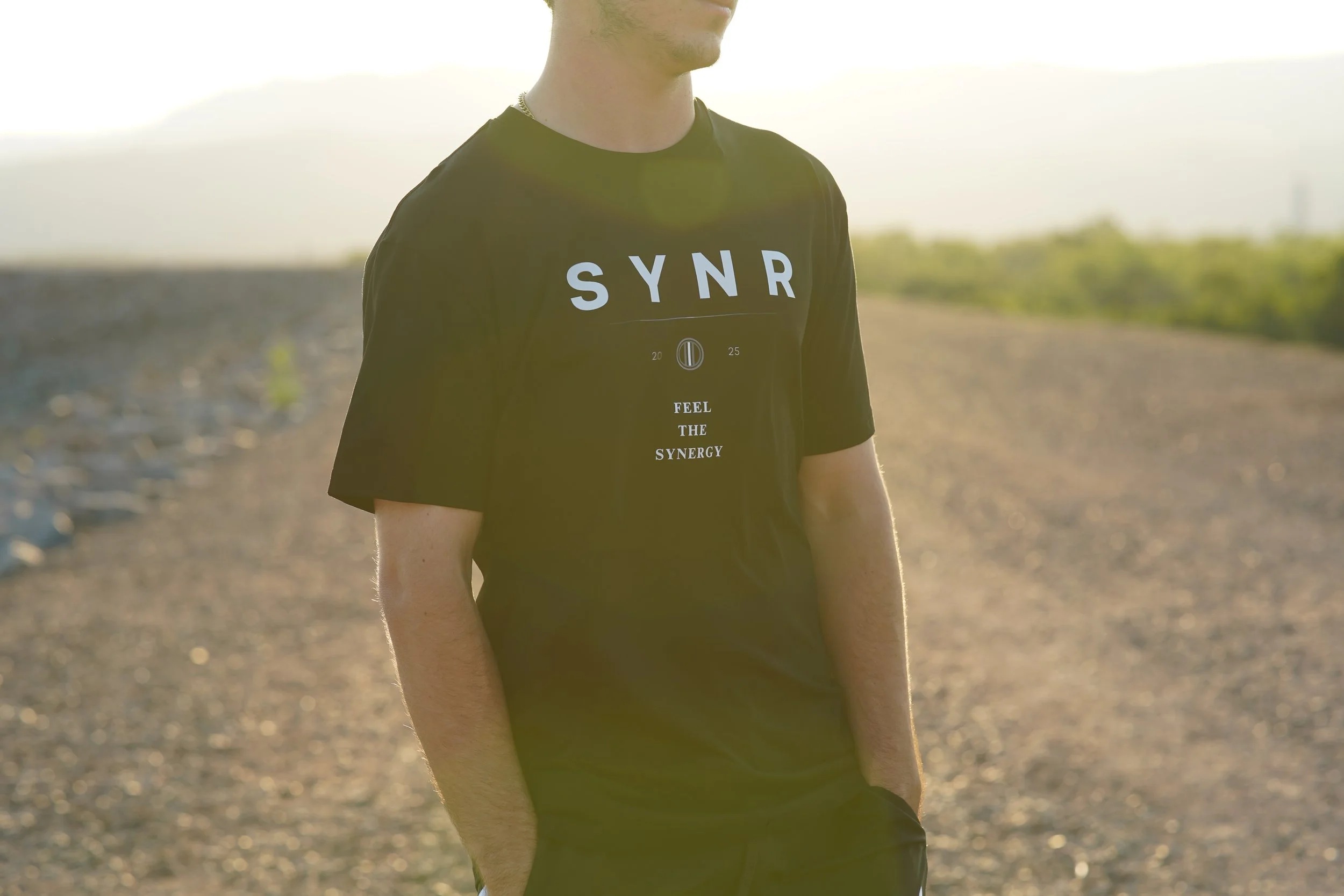 A person wearing a black t-shirt with white text standing outdoors in a rural area with a gravel road and green vegetation, sunlight in the background.