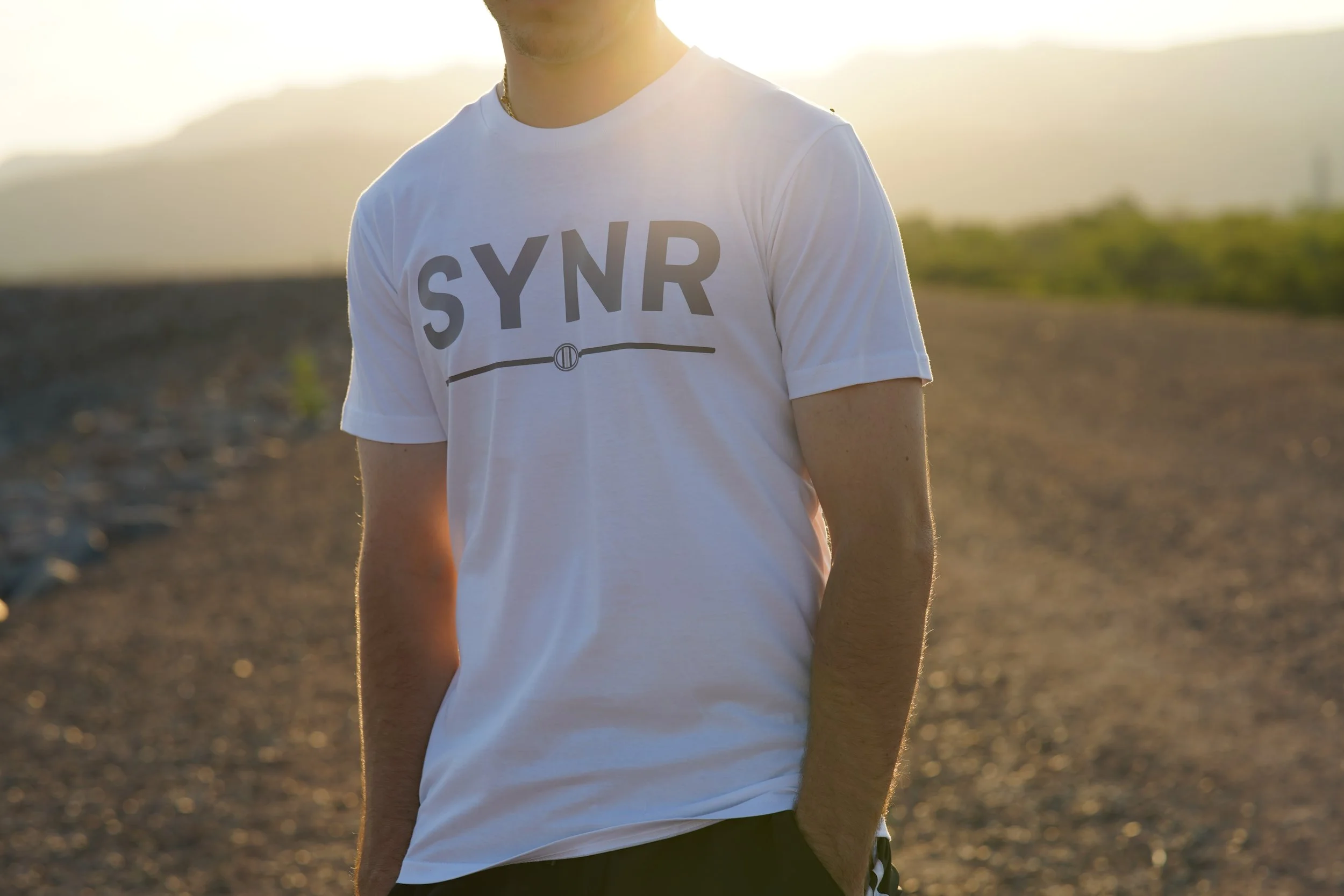 Close-up of a man wearing a white T-shirt with the word 'SYNR' printed on it, standing outdoors during sunset with a blurred landscape and mountains in the background.