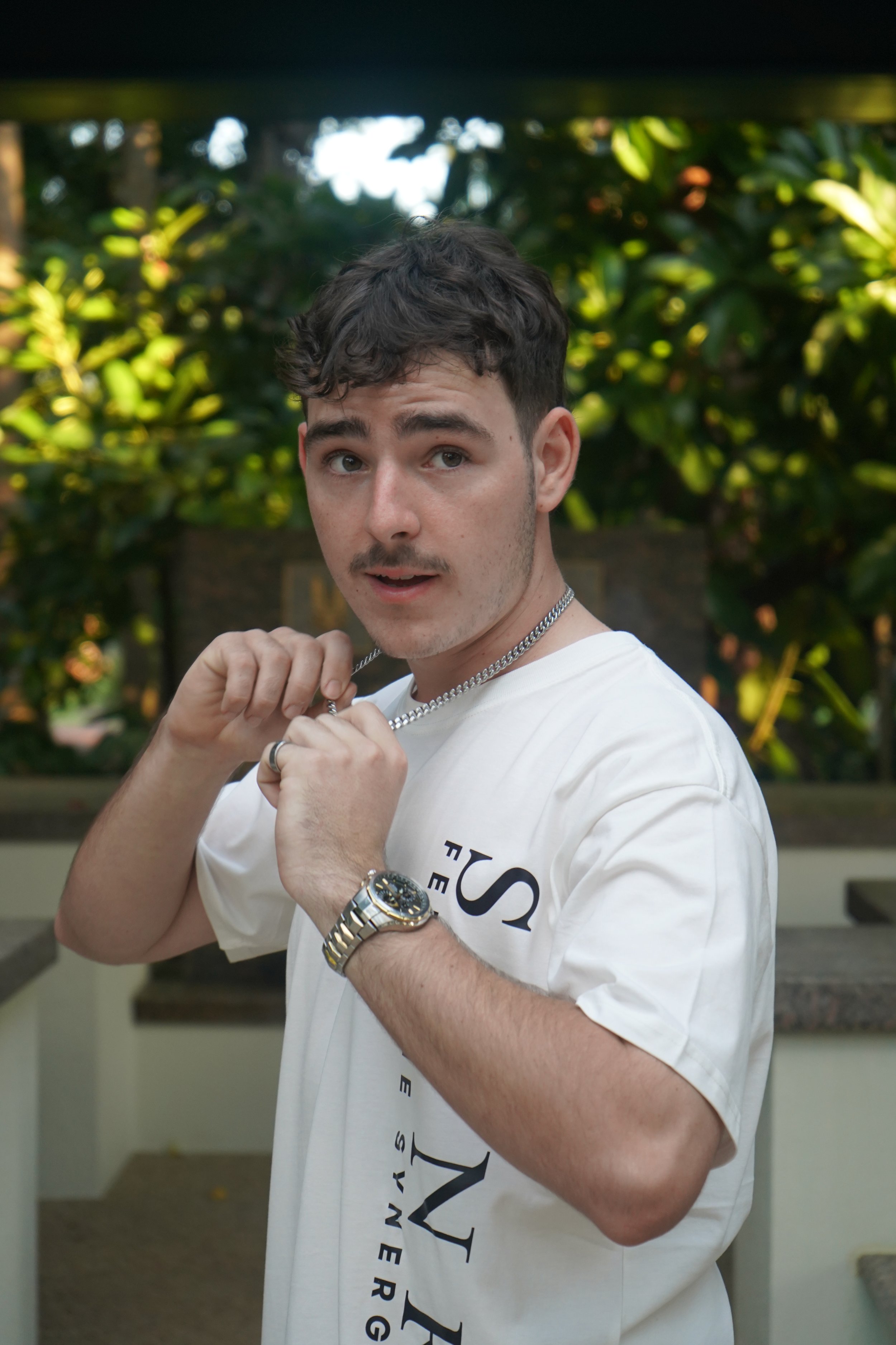 A young man with dark hair and light skin holding a chain necklace, wearing a white graphic T-shirt and a silver watch, standing outdoors with green foliage in the background.