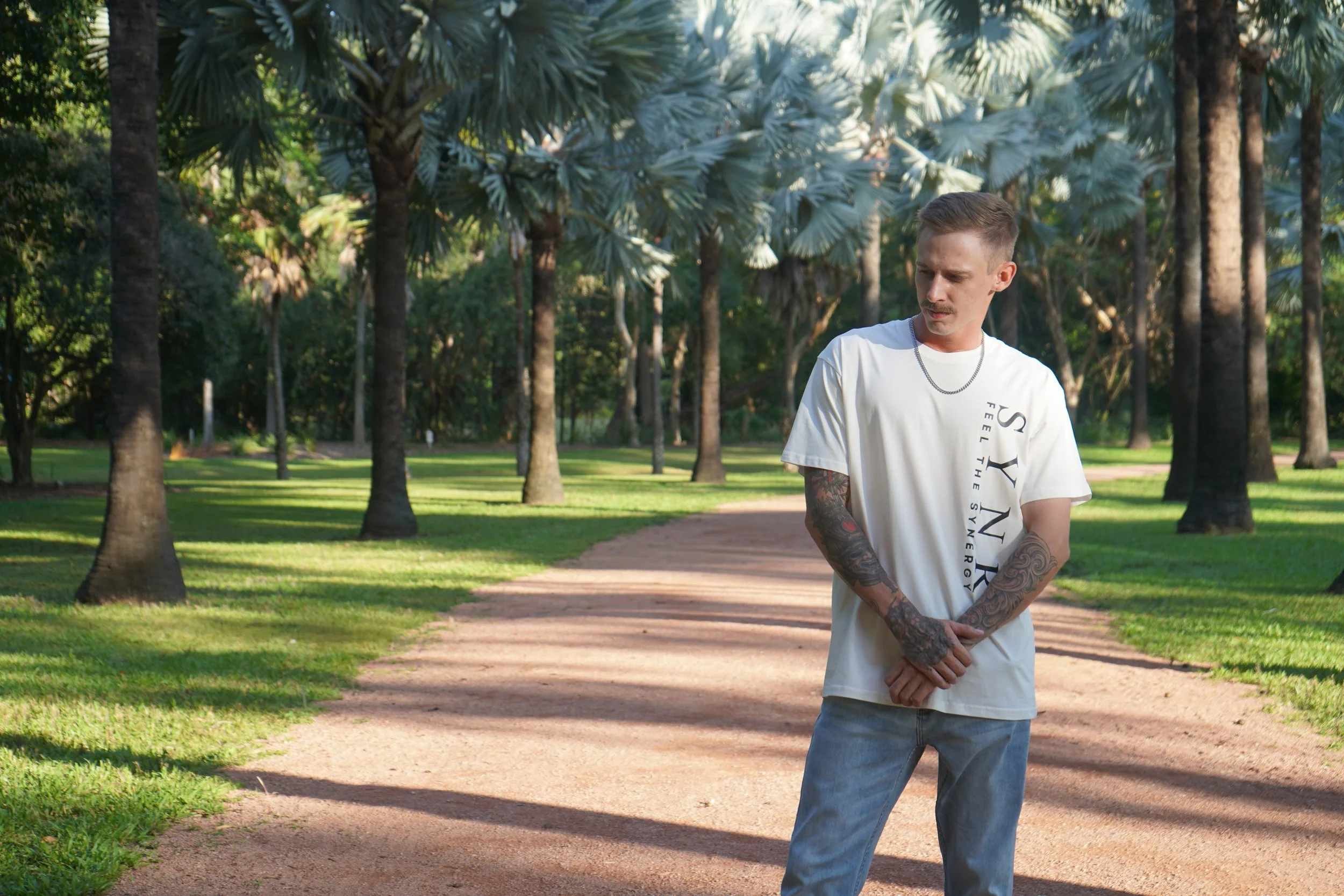 A young man with tattoos on his arms, wearing a white T-shirt and jeans, standing on a dirt path in a park with tall palm trees and green grass.