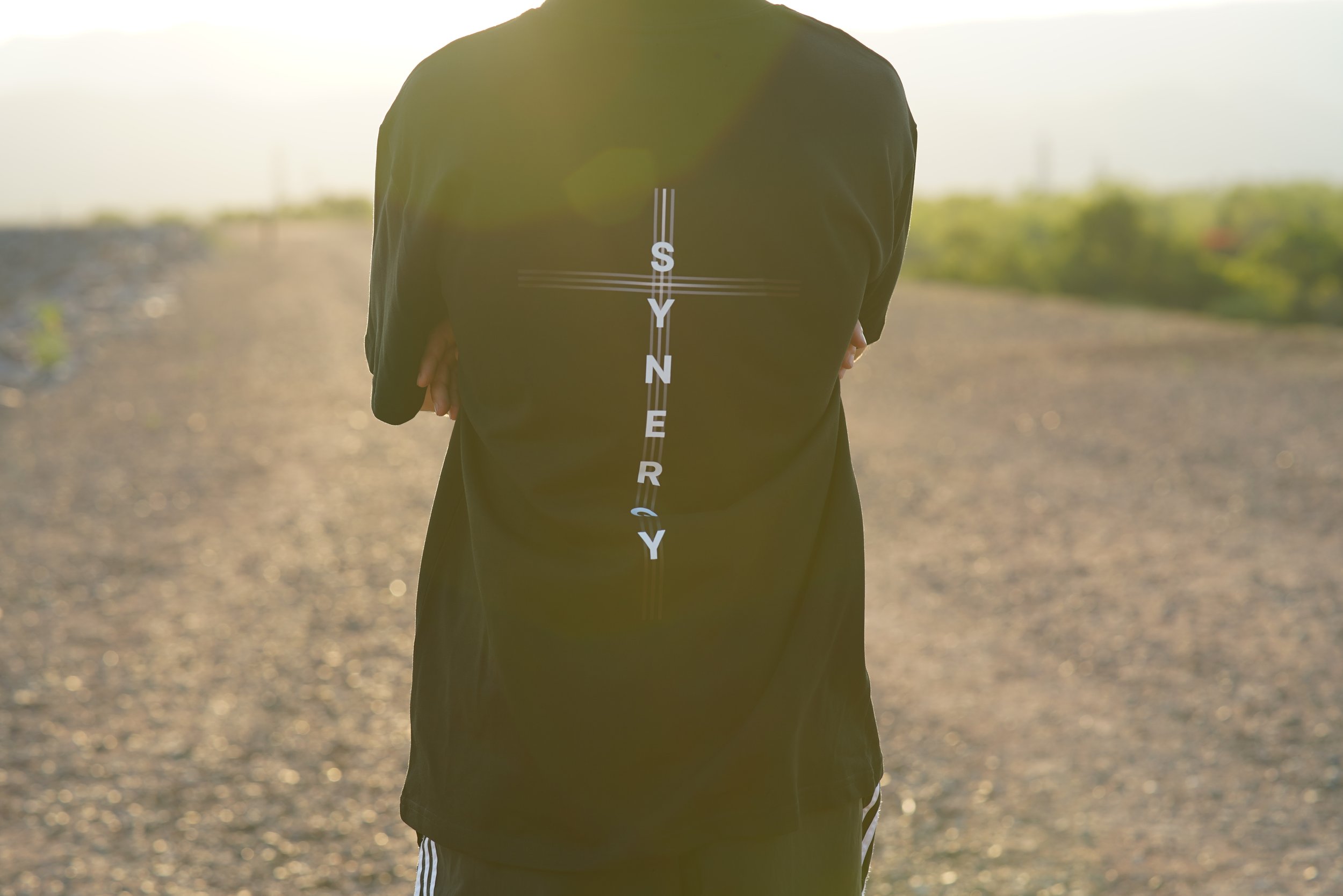 Person standing outdoors with arms crossed, wearing a black jacket with 'SYNERGY' written vertically on the back, on a gravel path with a blurred green landscape and bright sunlight.