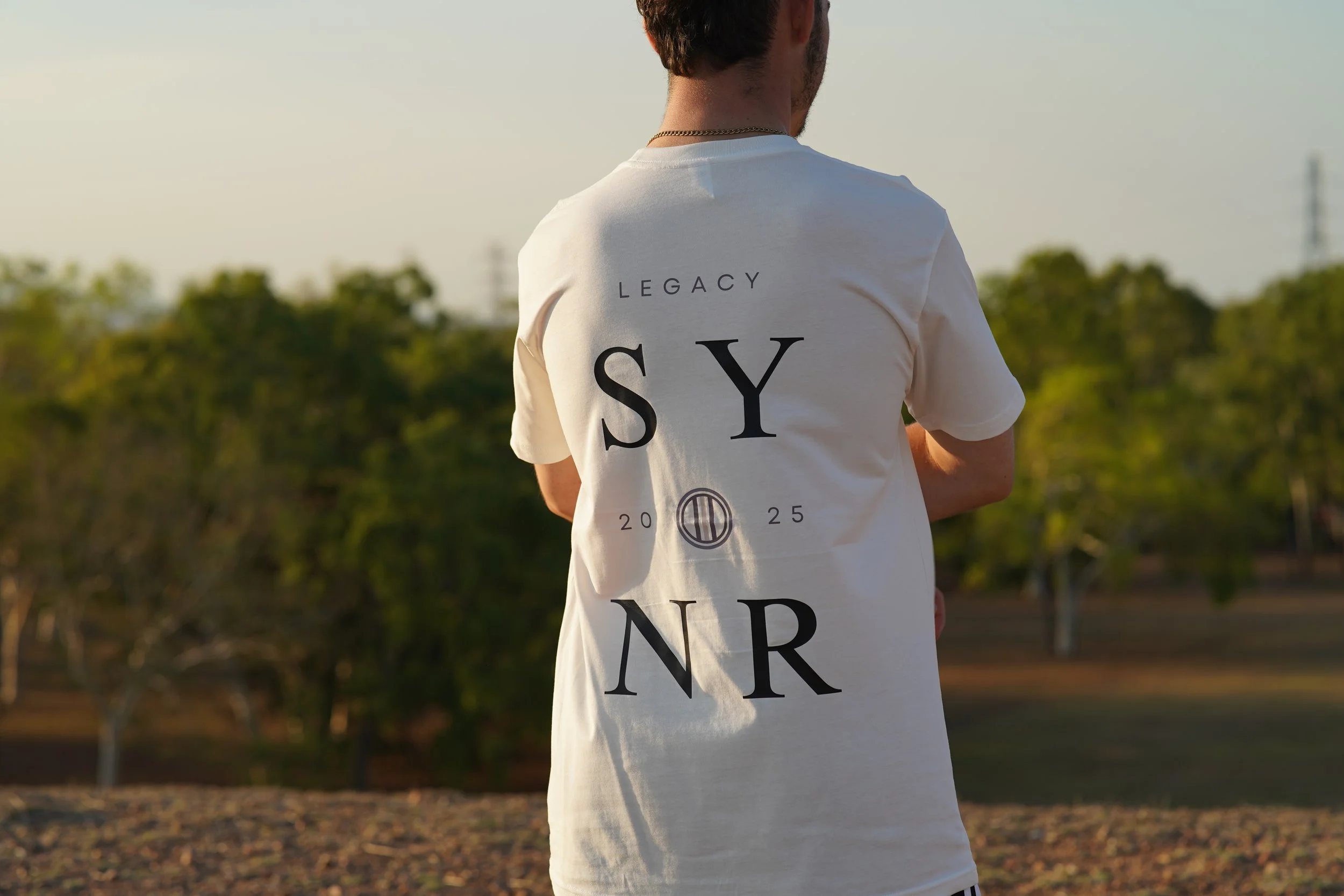 Back of a person wearing a white t-shirt with the word 'LEGACY' and 'SYNR' printed on it, standing outdoors during sunset with trees in the background.