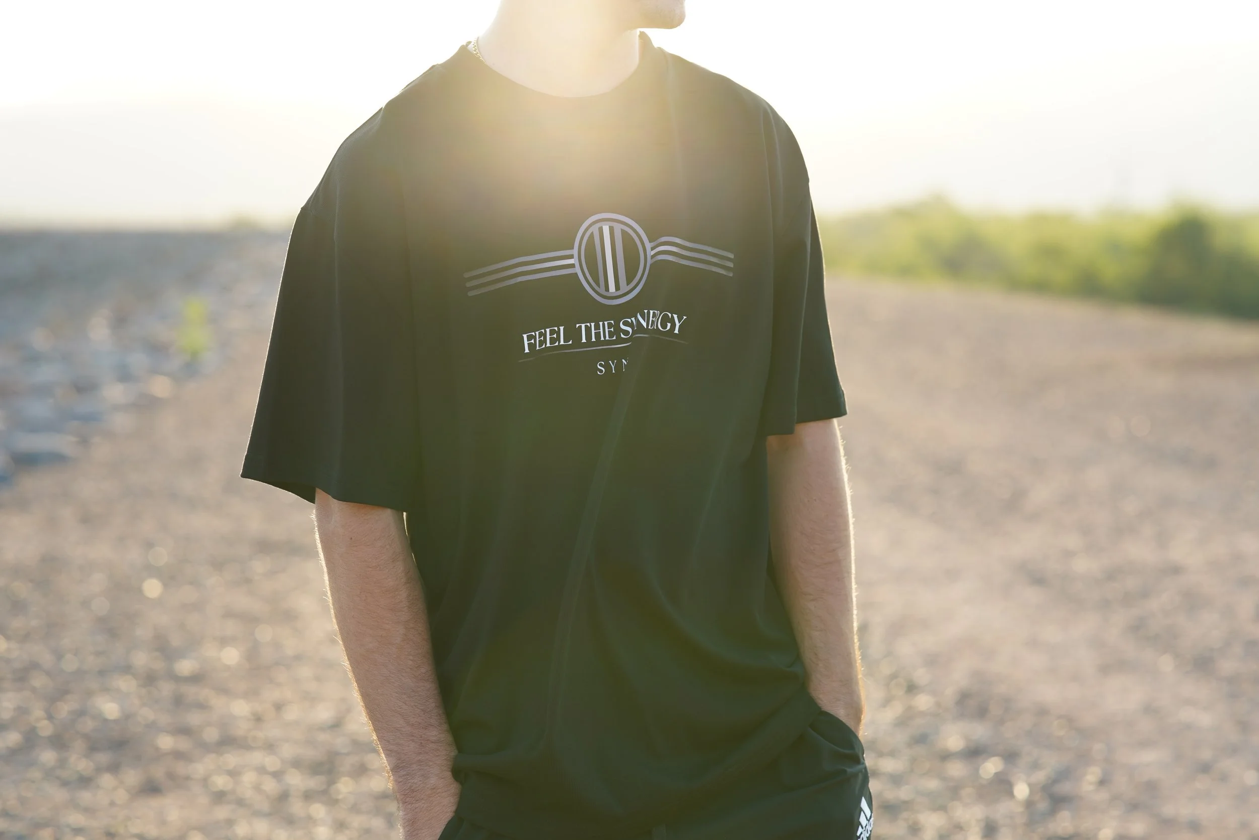 A man wearing a black T-shirt with the words "FEEL THE ENERGY" and a logo, standing outdoors on a gravel path with grass and trees in the distance, backlit by sunlight.