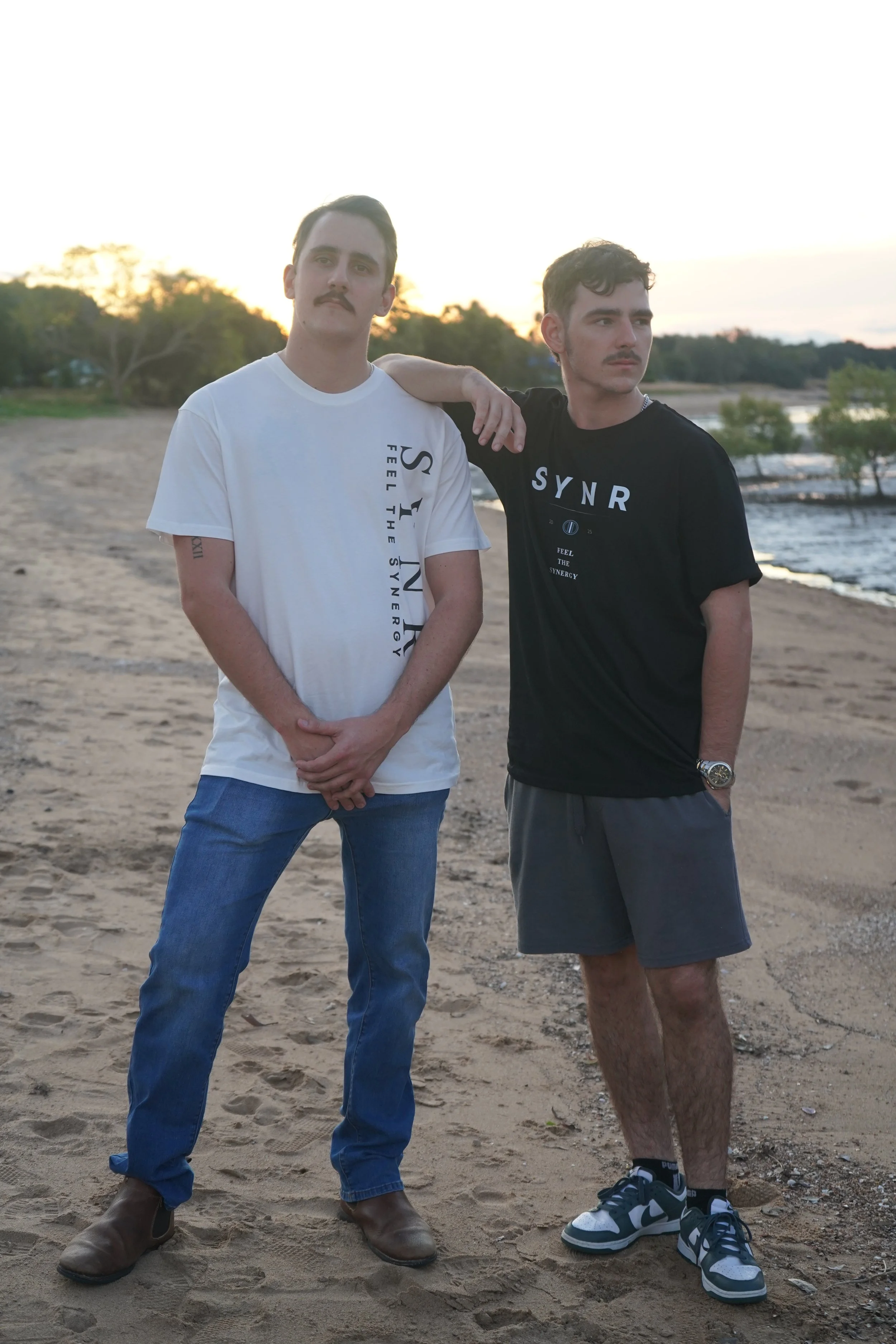 Two young men with mustaches standing on a sandy beach at sunset, one with arm around the other's shoulder, both wearing casual t-shirts and shorts.