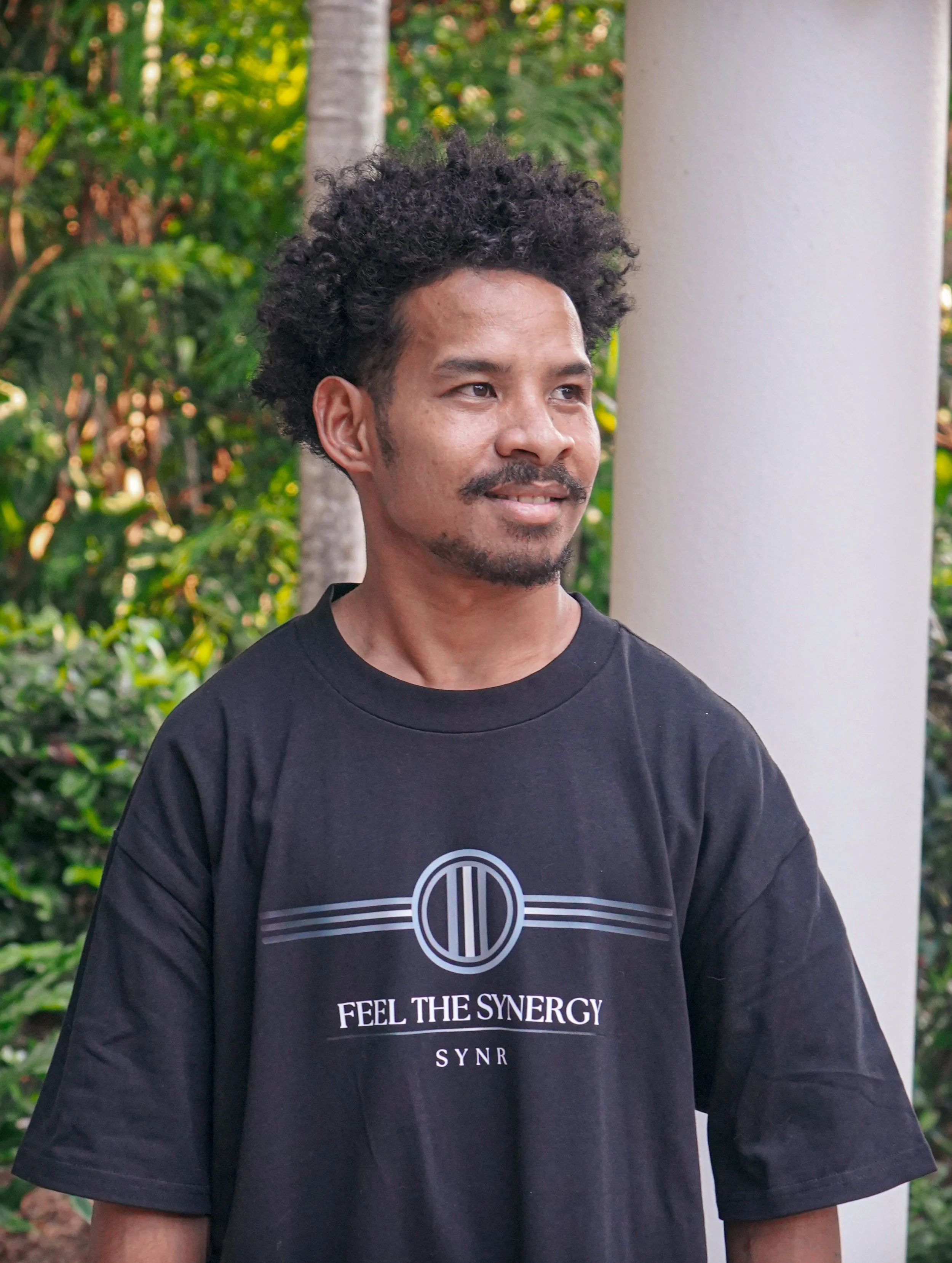 A man with curly black hair and facial hair standing outdoors near a white column, wearing a black t-shirt that says 'FEEL THE SYNERGY' across the chest, with green trees in the background.
