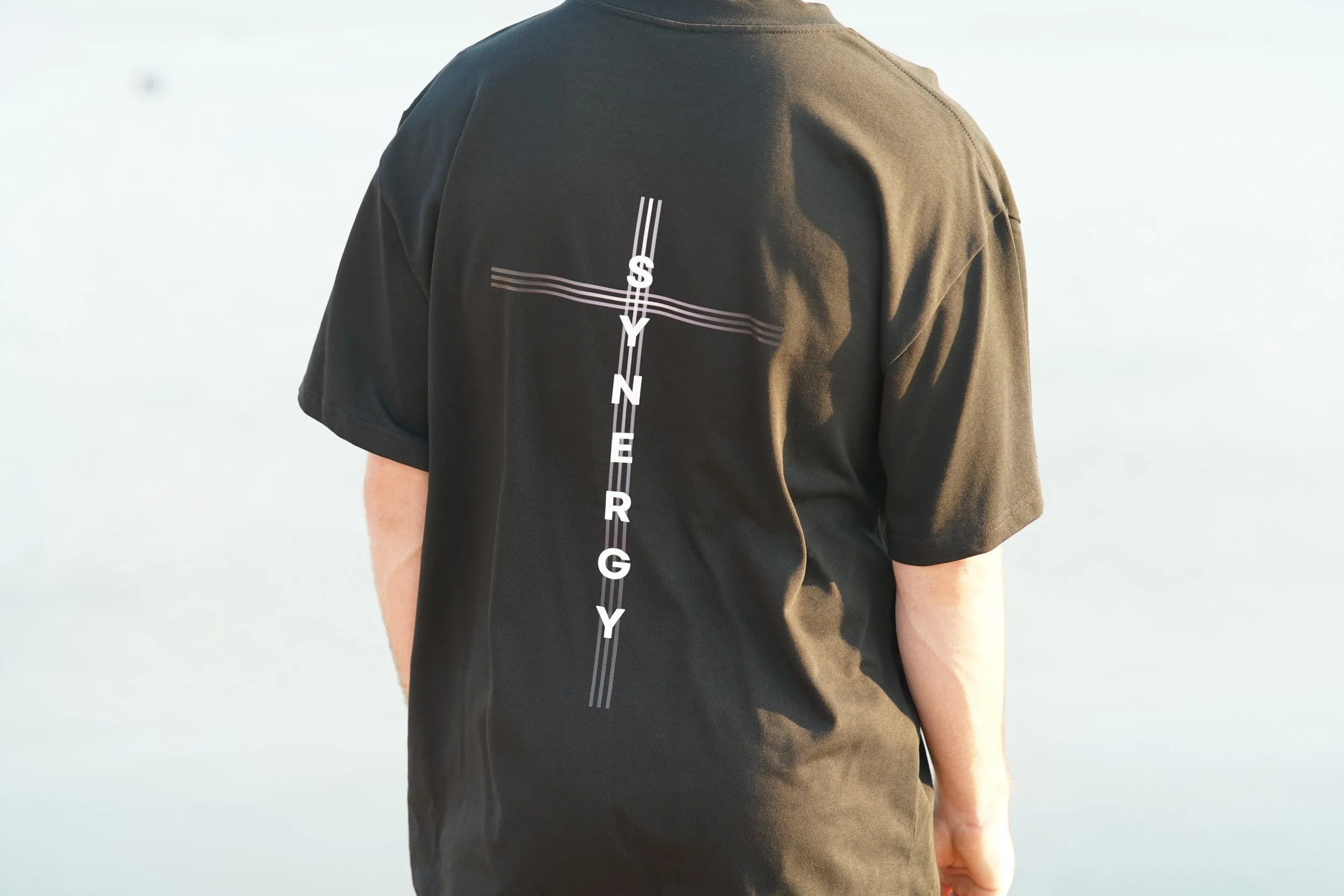 Person wearing a black T-shirt with the word 'SYNTHERY' written vertically along the back, and a cross with lines extending horizontally.