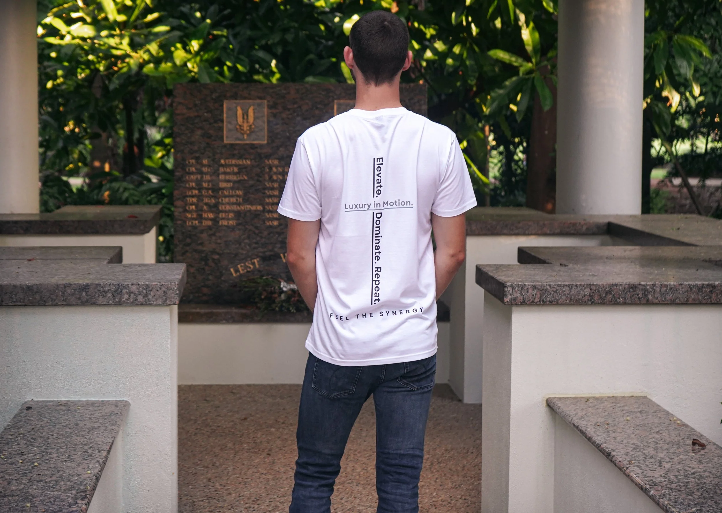 A person with short dark hair wearing a white T-shirt and dark jeans is standing with their back to the camera in front of a memorial plaque surrounded by lush green foliage.