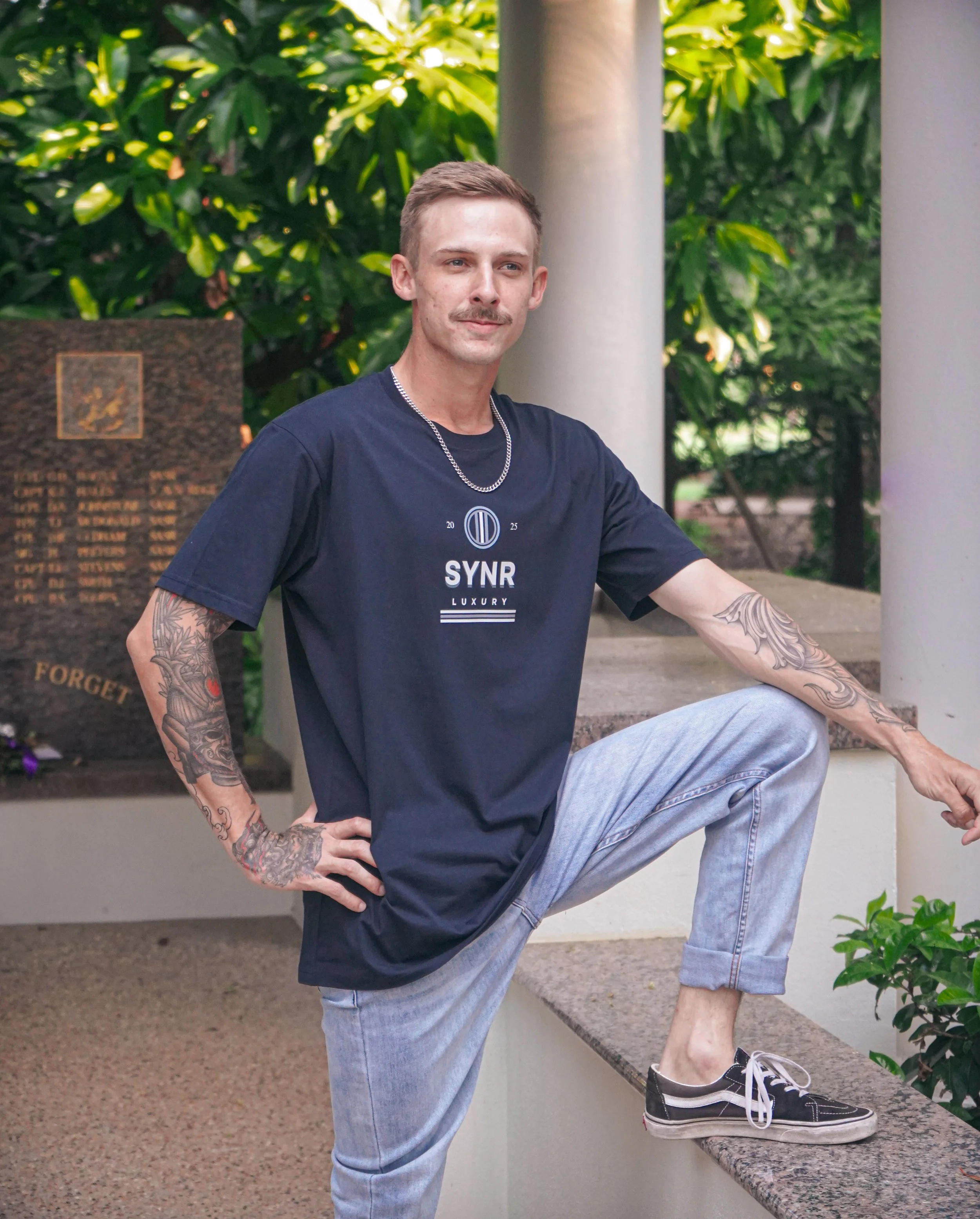 A young man with tattoos on his arms, wearing a black T-shirt with white text and a chain, blue jeans, and black sneakers, standing outdoors near a monument and greenery.