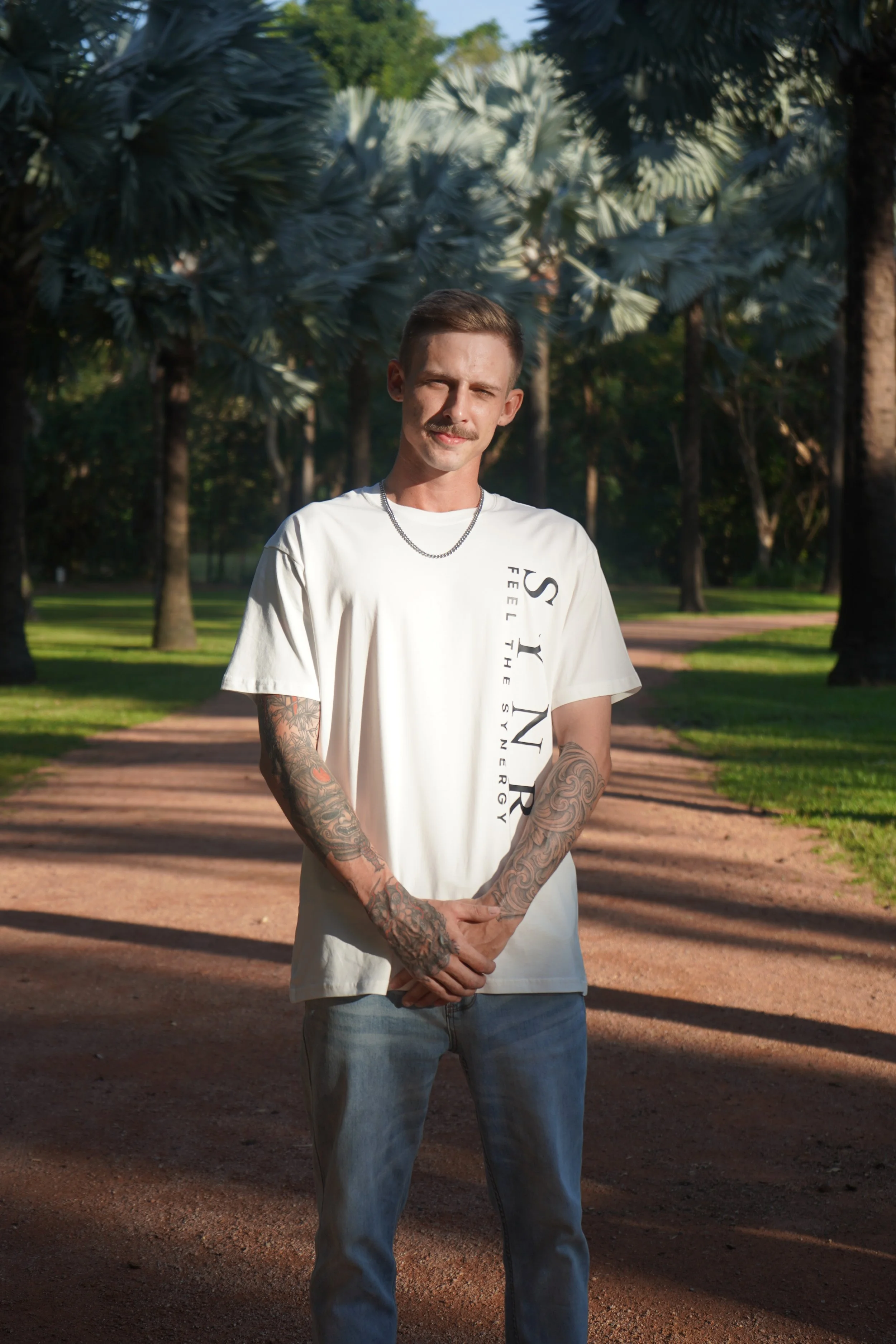 A young man with tattoos on his arms, wearing a white T-shirt with black text and light blue jeans, stands on a dirt path in a park with tall trees and lush greenery, during late sunlight.