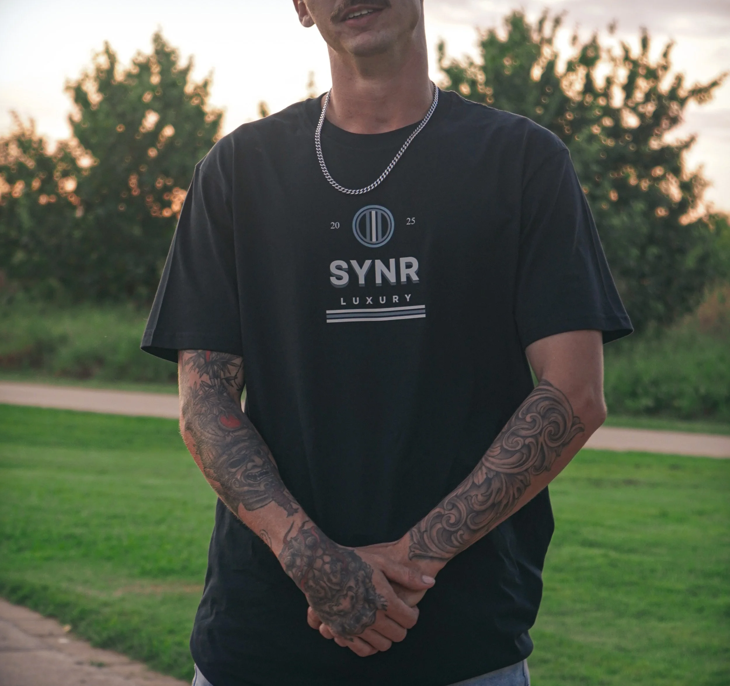 A man with tattoos on his arms wearing a black T-shirt with the words 'SYN R Luxury' and a chain necklace, standing outdoors at sunset or dusk, with grass and trees in the background.