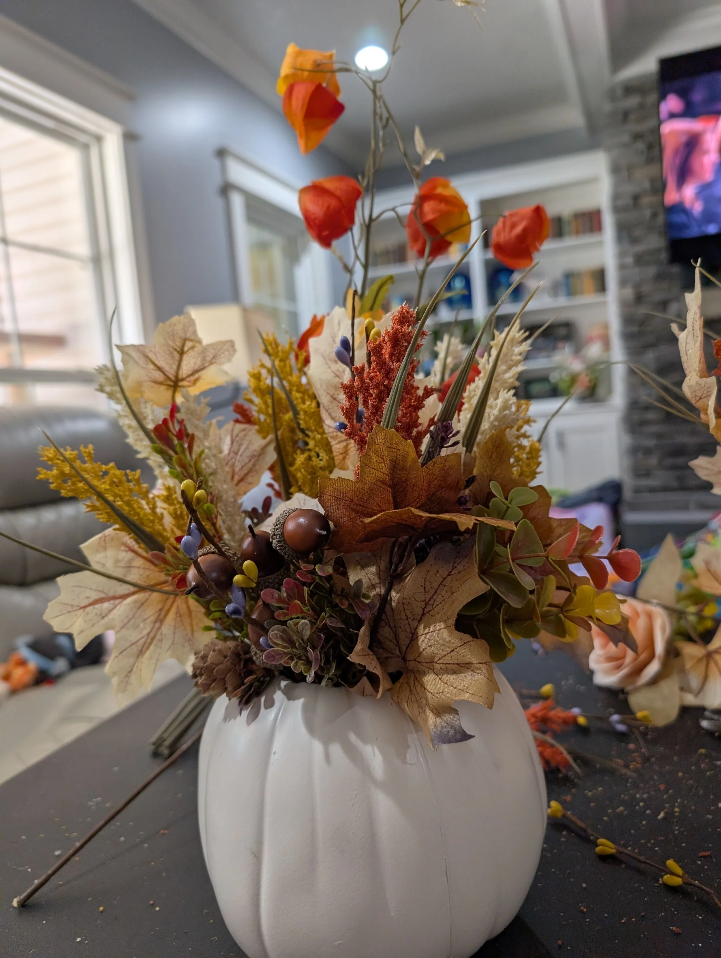 A white pumpkin arrangement with pale veined leaves, orange lantern-shaped accents, pinecones and acorn details.