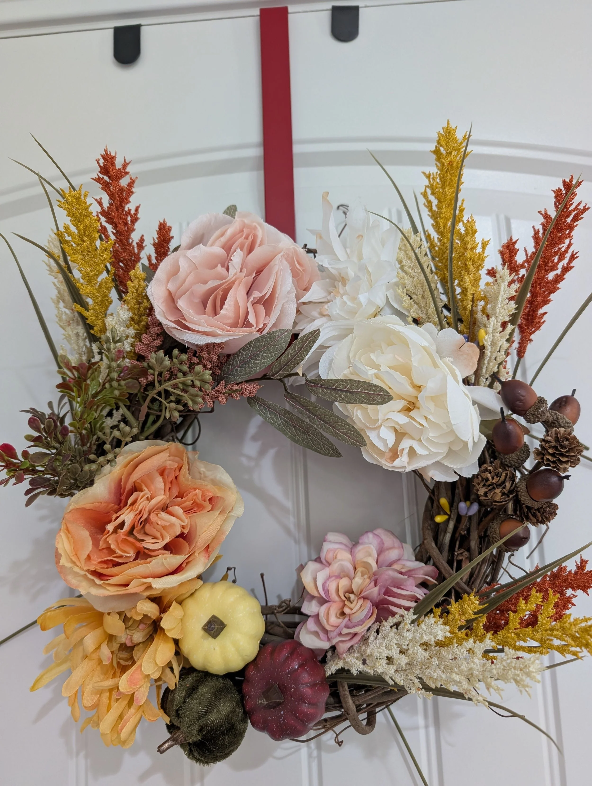 A floral wreath on a white door with blush, peach and cream flowers, small decorative pumpkins, pinecones and golden stems.