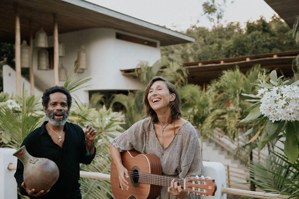Two people outdoors, one playing guitar and the other holding a vase, surrounded by lush plants and white flowers, with a modern building in the background, smiling and enjoying themselves.