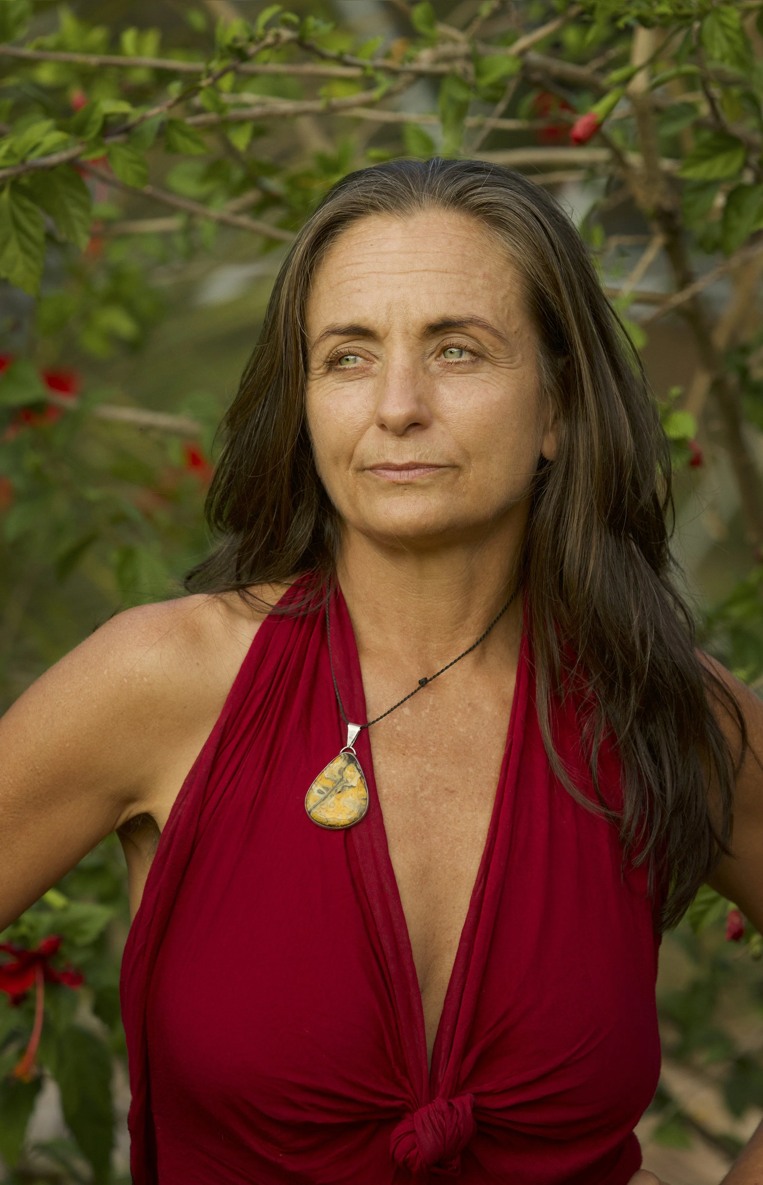 Una mujer con el pelo largo y oscuro, vestida con un top rojo sin mangas y un collar con colgante amarillo, de pie al aire libre entre el follaje verde.