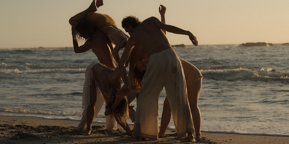 Un grupo de cinco personas está jugando y bailando en la playa al atardecer o al amanecer, cerca del agua del mar.