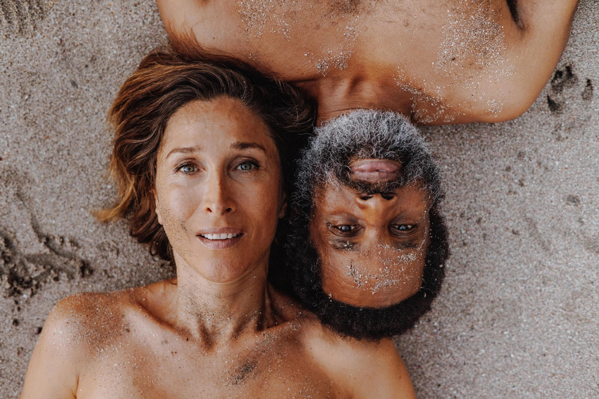 A couple with sandy skin lying on a sandy beach, facing the camera and resting their heads close together.