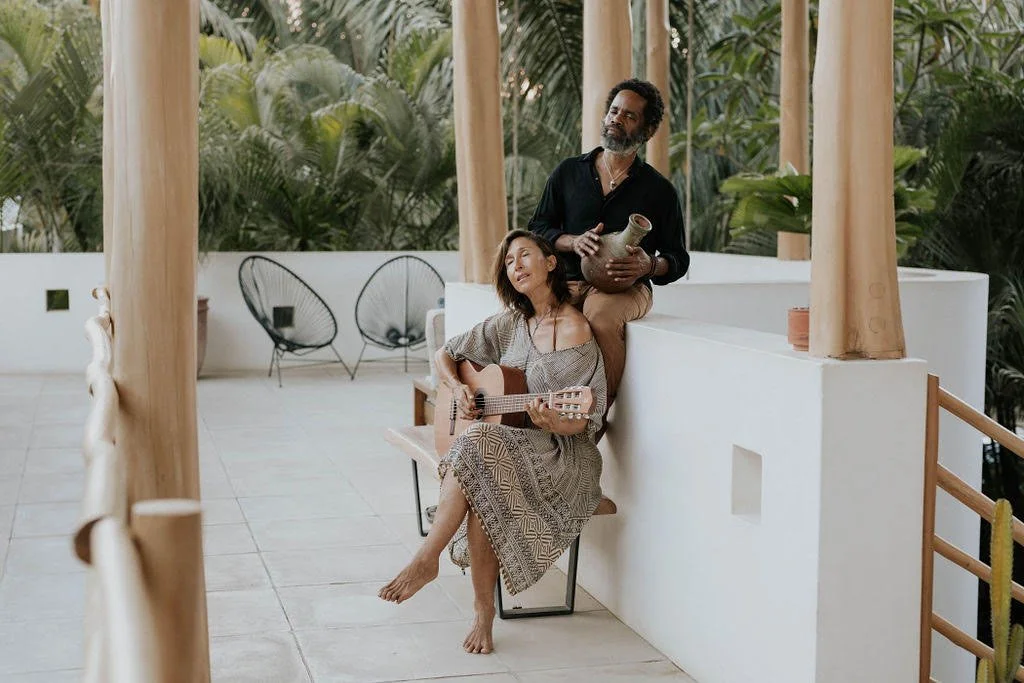 A woman sitting on a stool playing guitar and singing, a man sitting behind her holding a ceramic jug, on a balcony with plants and outdoor furniture.