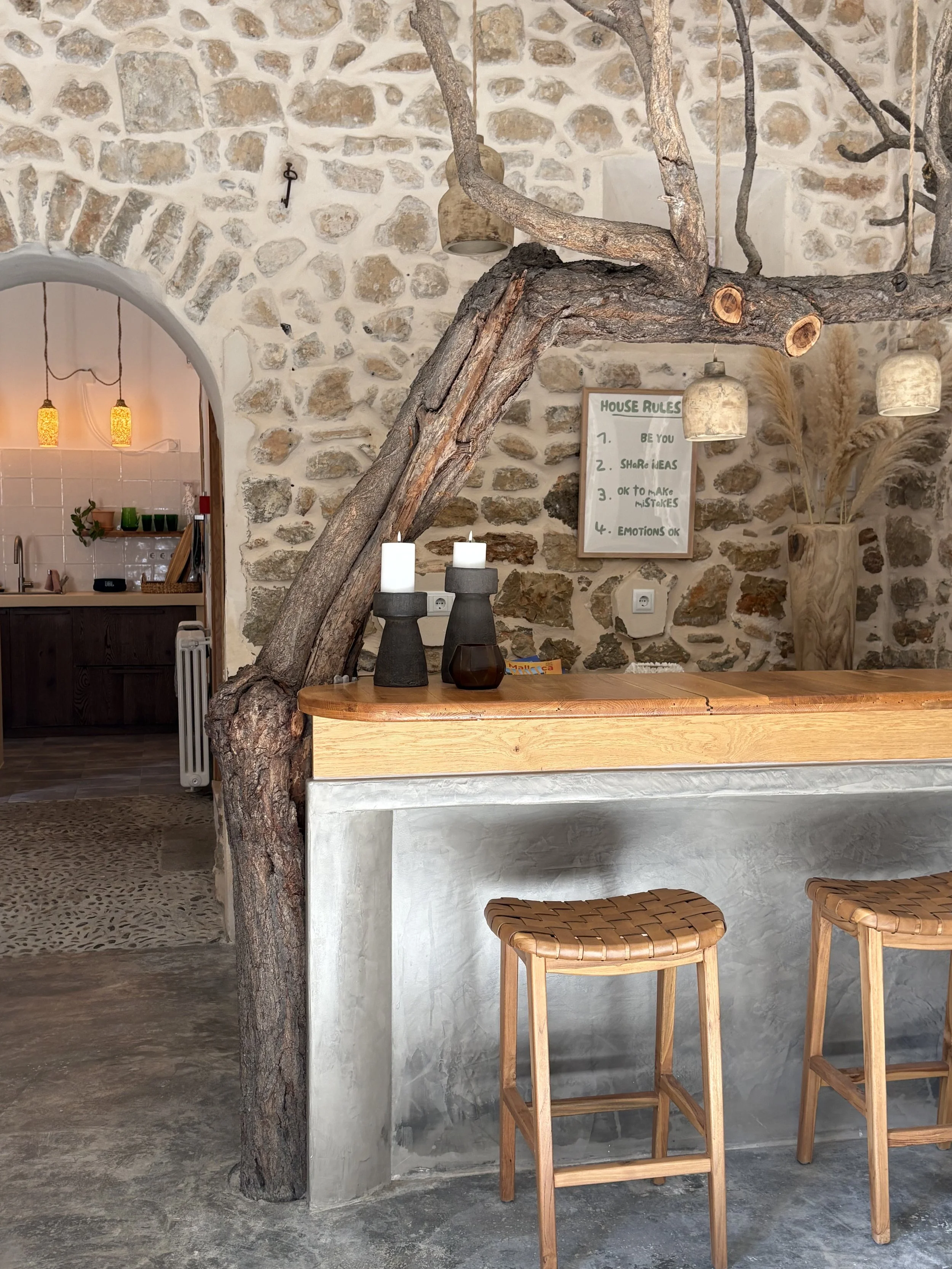 Interior of a rustic room with a stone wall, a wooden bar countertop, two woven wooden stools, candles in black holders, and house rules written on a framed board on the wall.