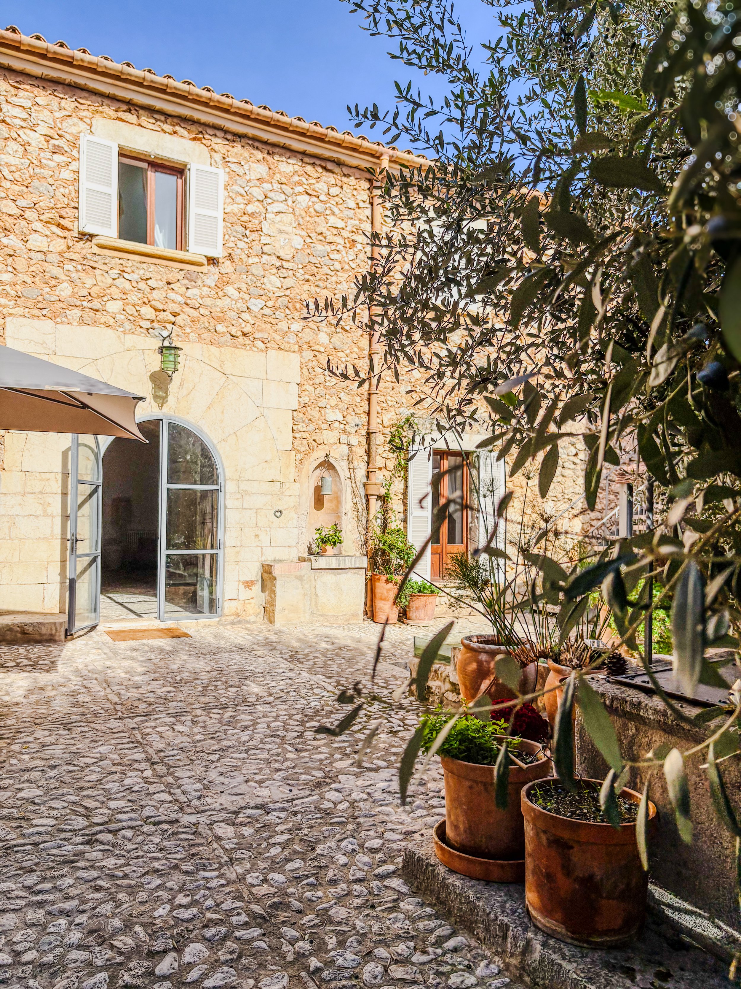 A sunny courtyard with a stone cobblestone ground, terracotta pots with plants, a stone and brick building with windows and open doors, an umbrella, and a tree with green leaves.