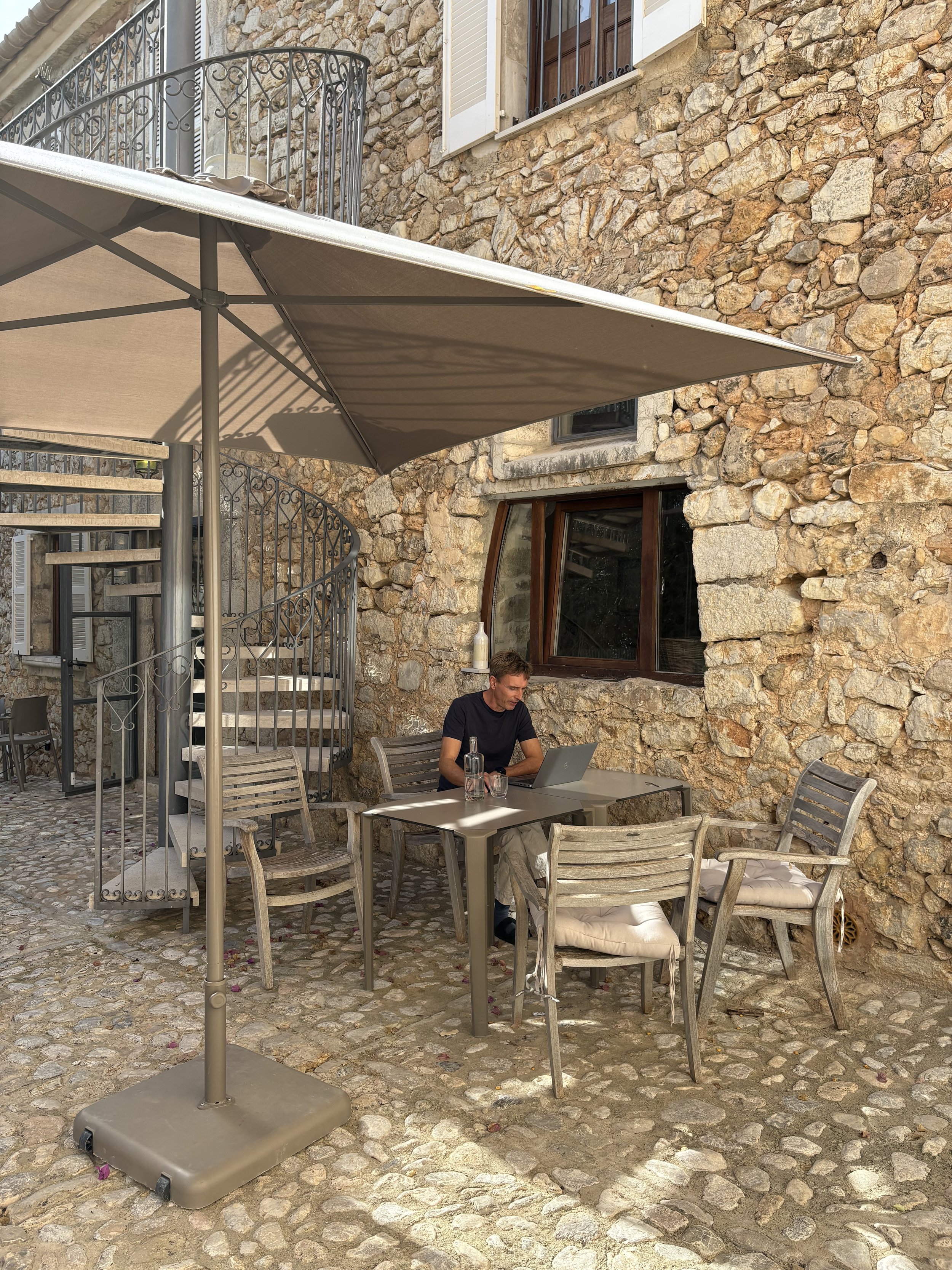 A man sitting at an outdoor table with a laptop and bottled water on a stone patio, shaded by a large umbrella, next to a stone wall with windows and a metal spiral staircase. He works remote and on his business with likeminded people for New Work
