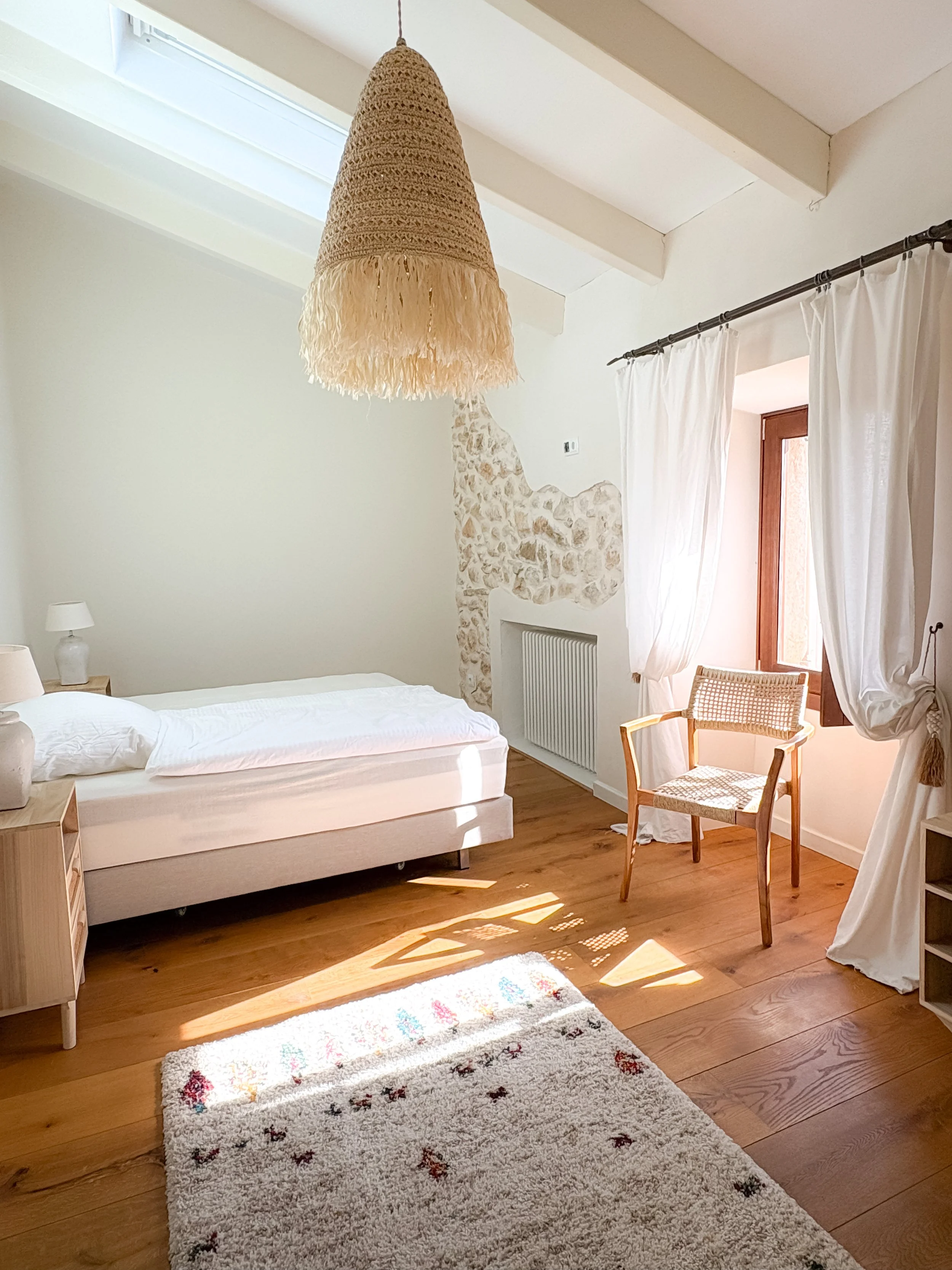 Sunlit bedroom with a single bed, a bedside table with a lamp, a wooden chair near a window with white curtains, a woven lampshade hanging from the ceiling, a beige and red patterned rug, and a stone accent wall.