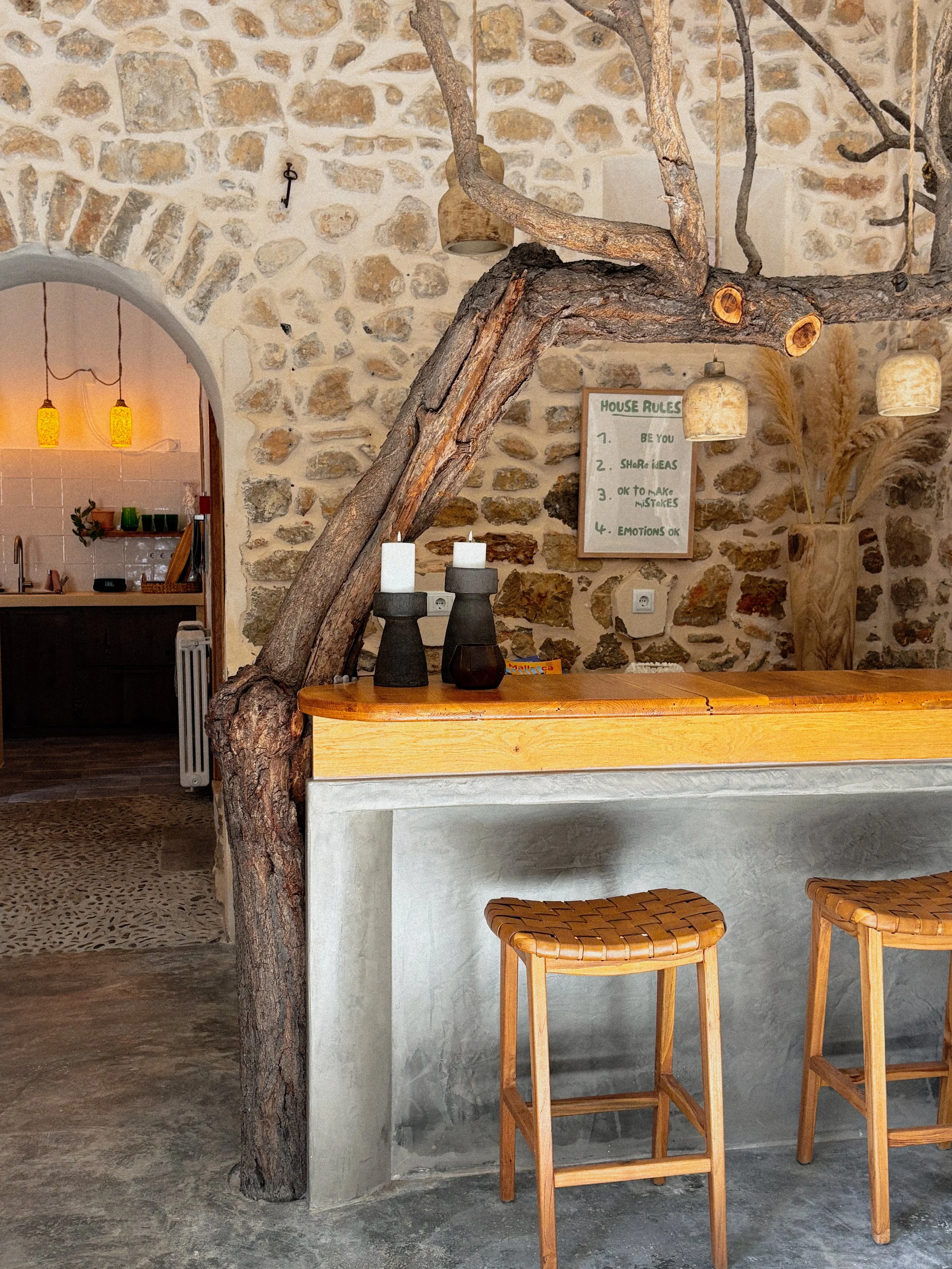 Interior of a rustic-style kitchen and dining area featuring a stone wall, wooden bar with two bar stools, decorative branches overhead, and a house rules sign on the wall.