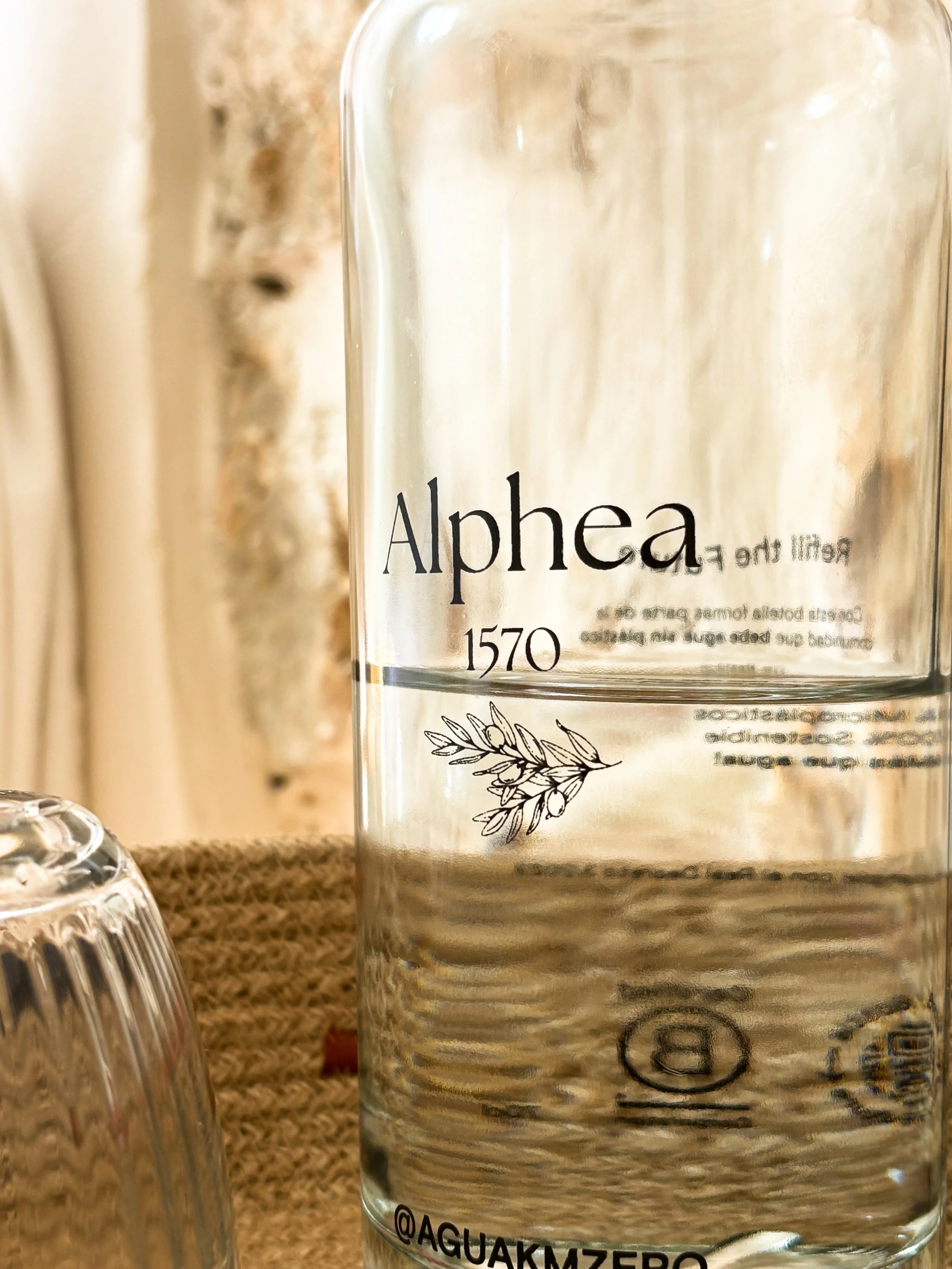 Close-up of a transparent glass bottle labeled "Alphea 1570" with a simple black illustration of an olive branch, placed on a textured surface with a blurry background.