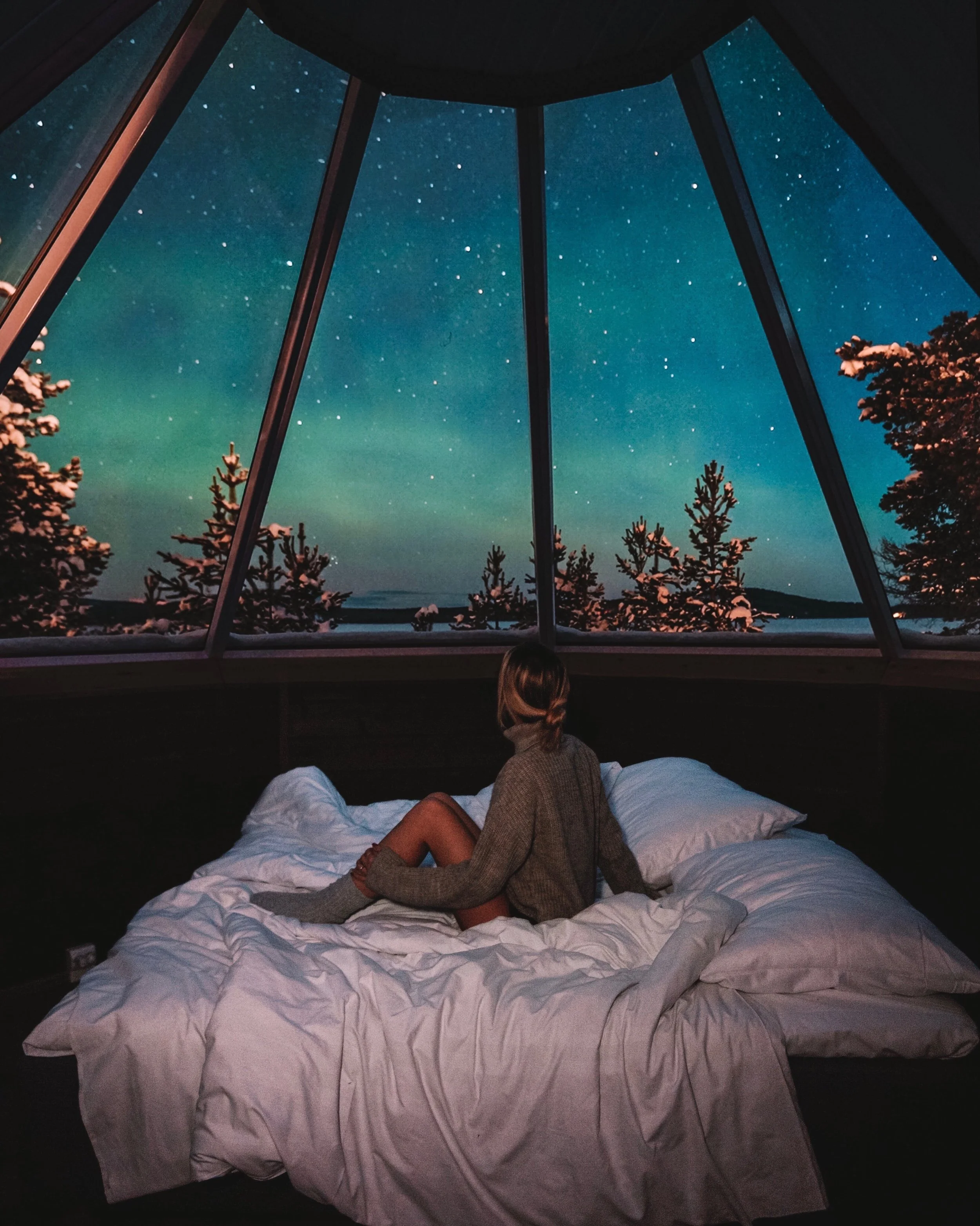 Woman sitting on bed in a cozy room, gazing out a large panoramic window at a starry night sky and shooting stars over a forested landscape.