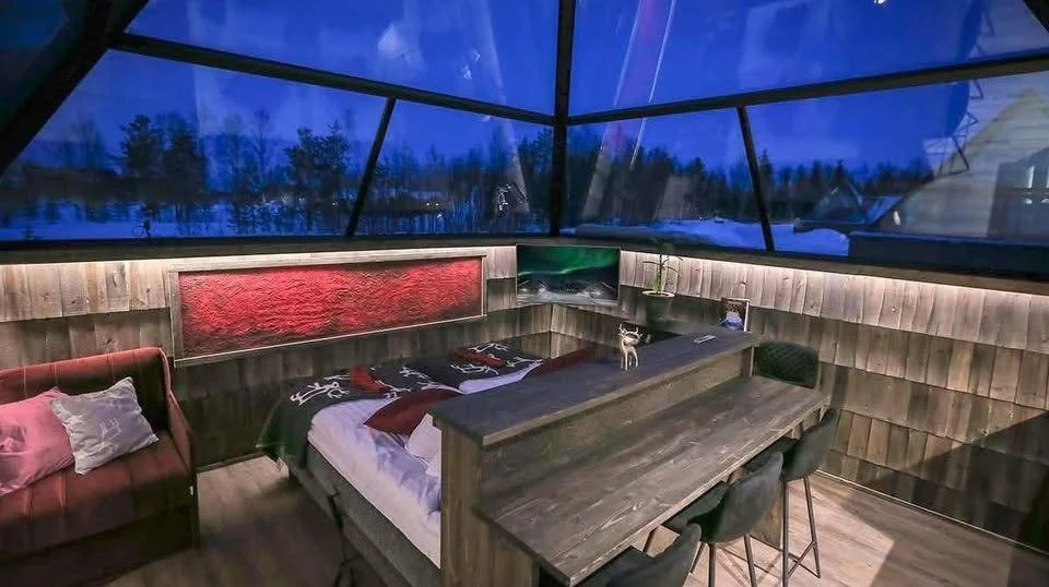 Interior of a cozy, modern cabin with a large window showcasing a snowy winter landscape outside. The room features wooden paneling, a bed with red and white bedding, a small red sofa with pillows, a dining table with four chairs, and decorative pill