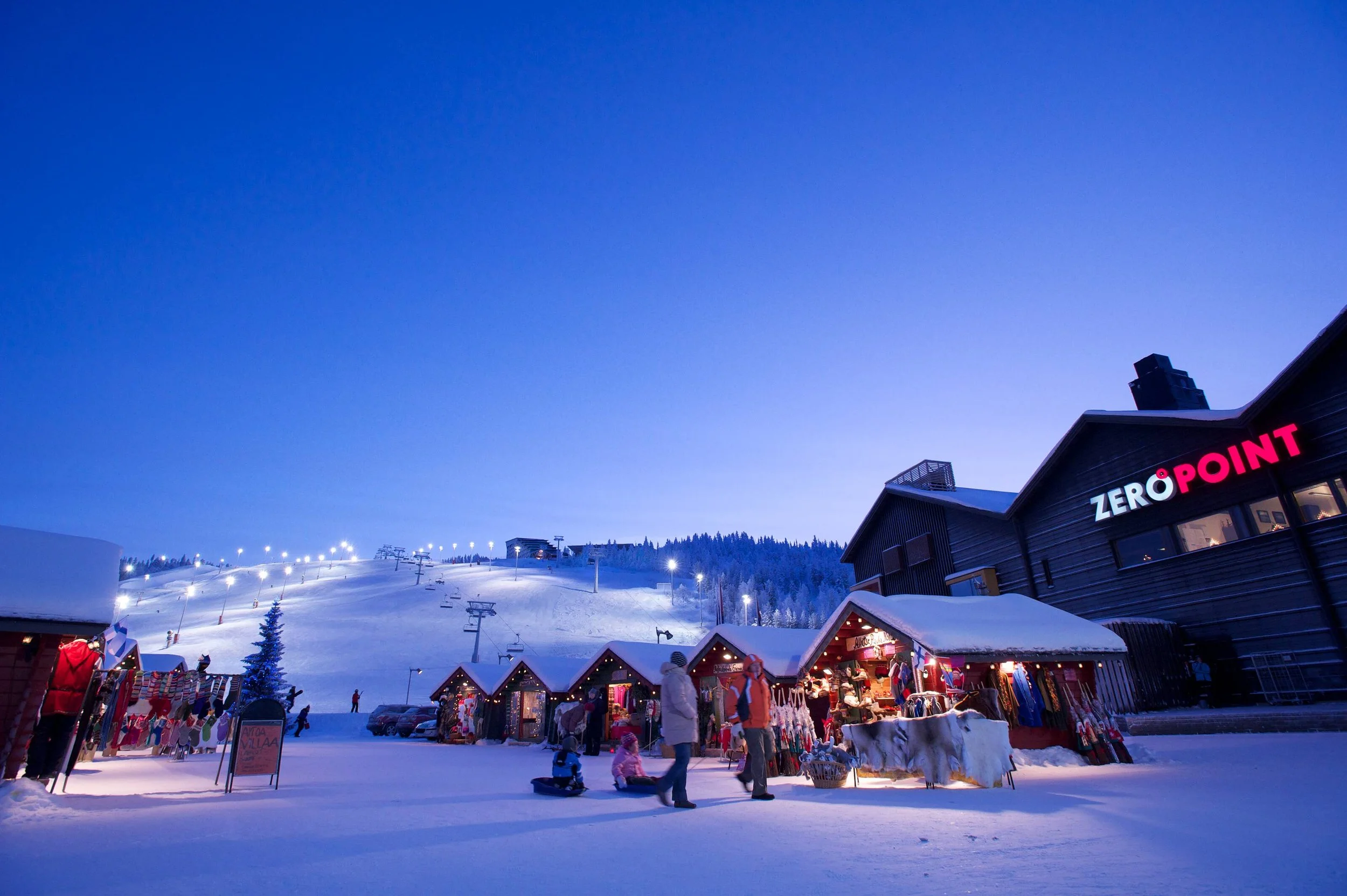 Snow-covered outdoor market at dusk with small wooden stalls, people shopping, and ski slopes in the background under a blue sky. The building has a sign reading 'Zero Point'.