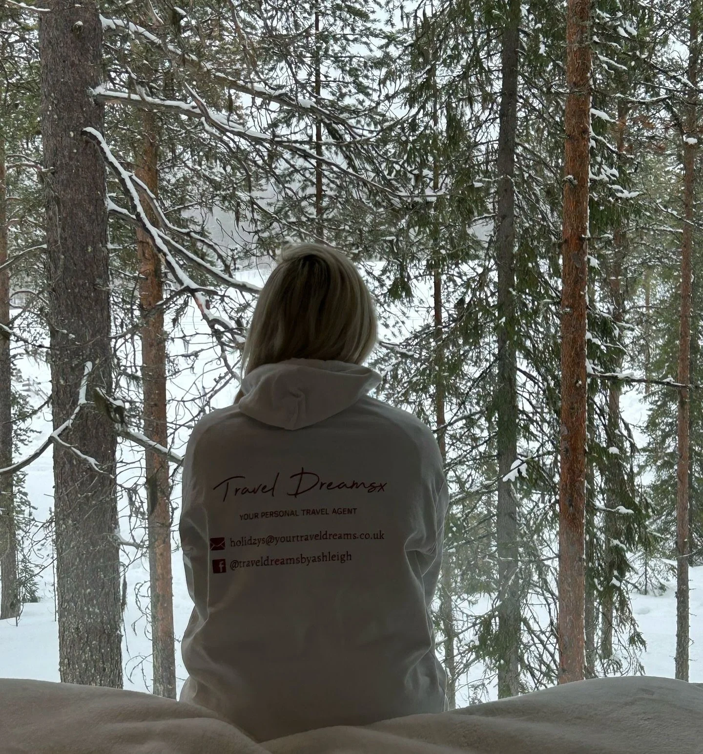 A woman with blond hair wearing a white hoodie sits on a bed facing a large window, looking out at a snowy forest with tall trees and snow-covered branches.