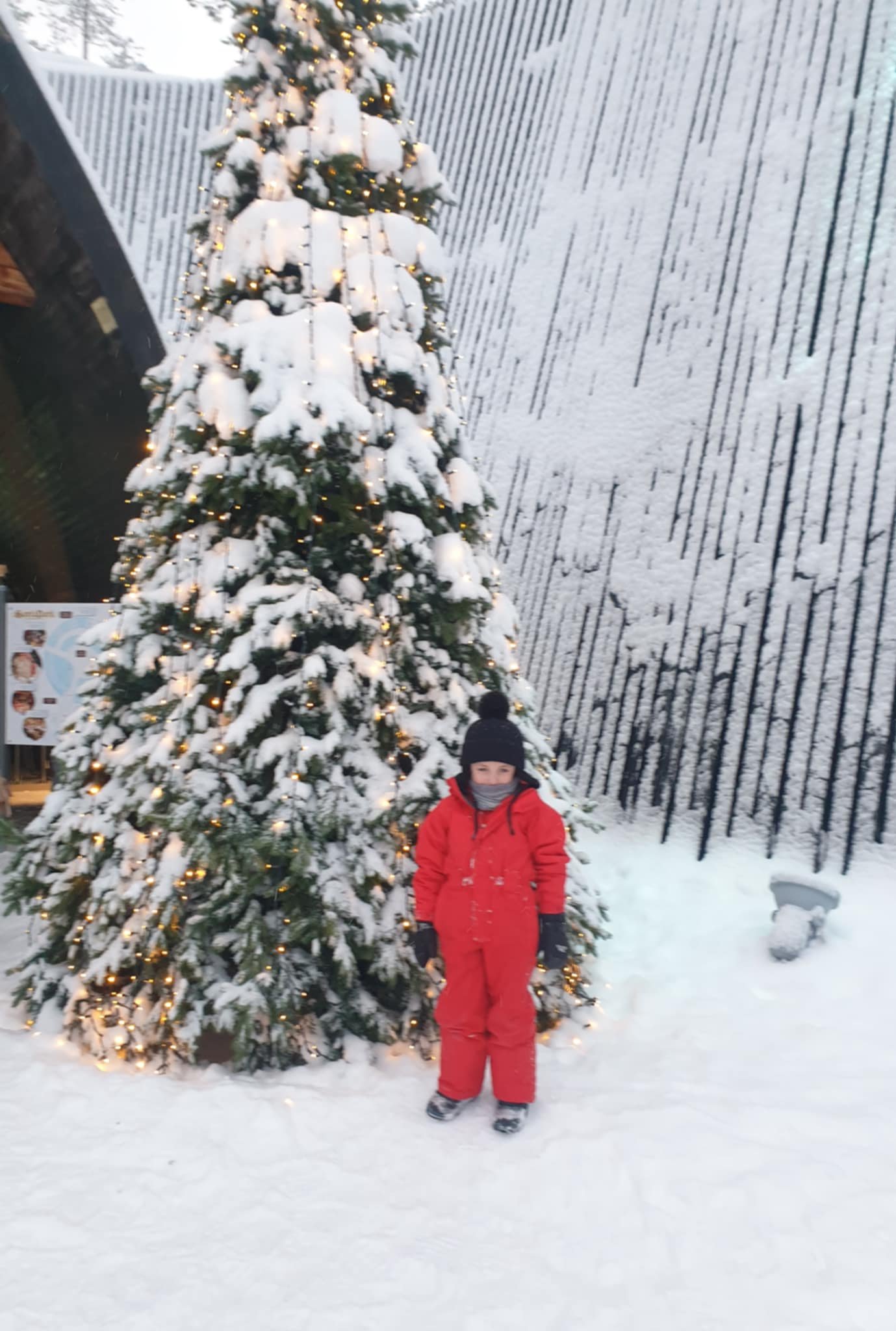 A child in a red snowsuit, black gloves, and a black knit hat with a pom-pom stands in front of a snow-covered Christmas tree decorated with white lights. Snow is on the ground and on the tree, with a wooden wall in the background.