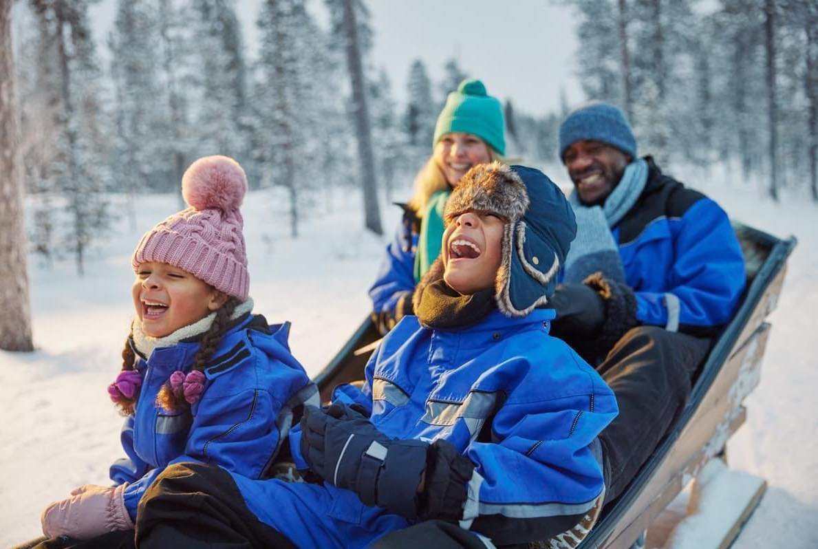 A family of four enjoying a winter moment outdoors, sitting on a sled in a snowy forest, dressed in colorful winter gear and smiling.