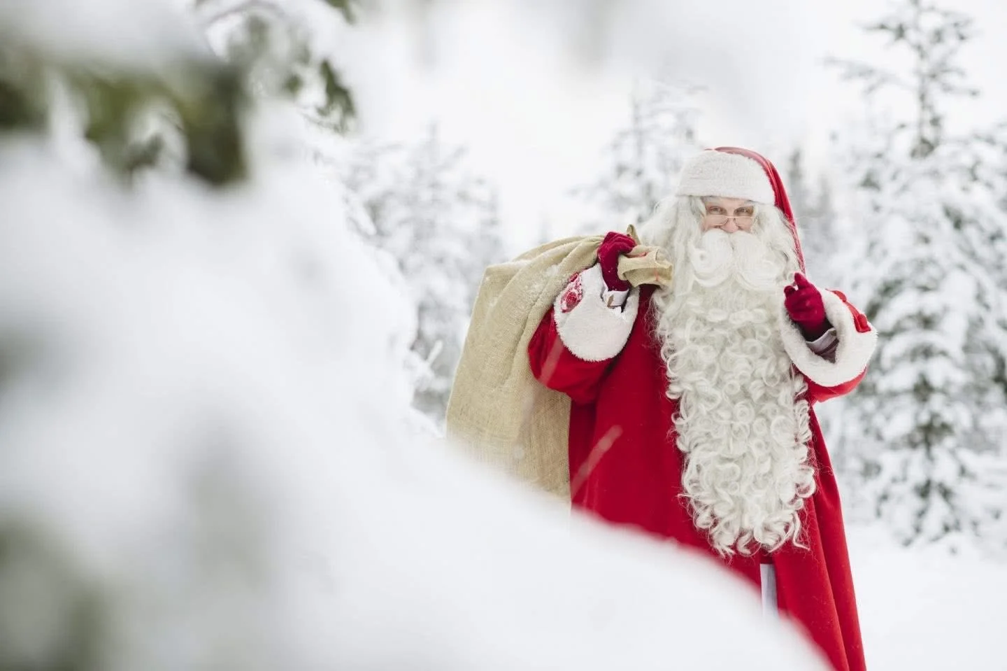 Santa Claus in a snowy forest carrying a burlap sack over his shoulder, dressed in a red suit with white fur trim, red gloves, and a red hat with white pom-pom.