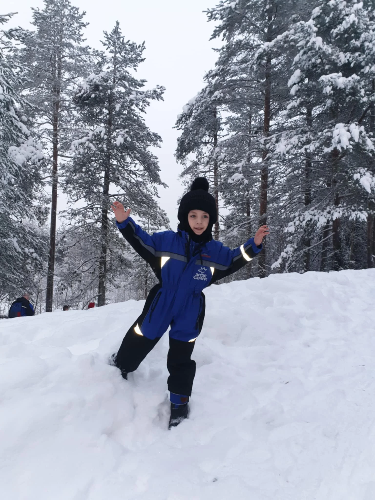 A young boy wearing a blue and black snowsuit and a black hat with a pom-pom is standing in deep snow in a snowy forest, smiling with arms raised.