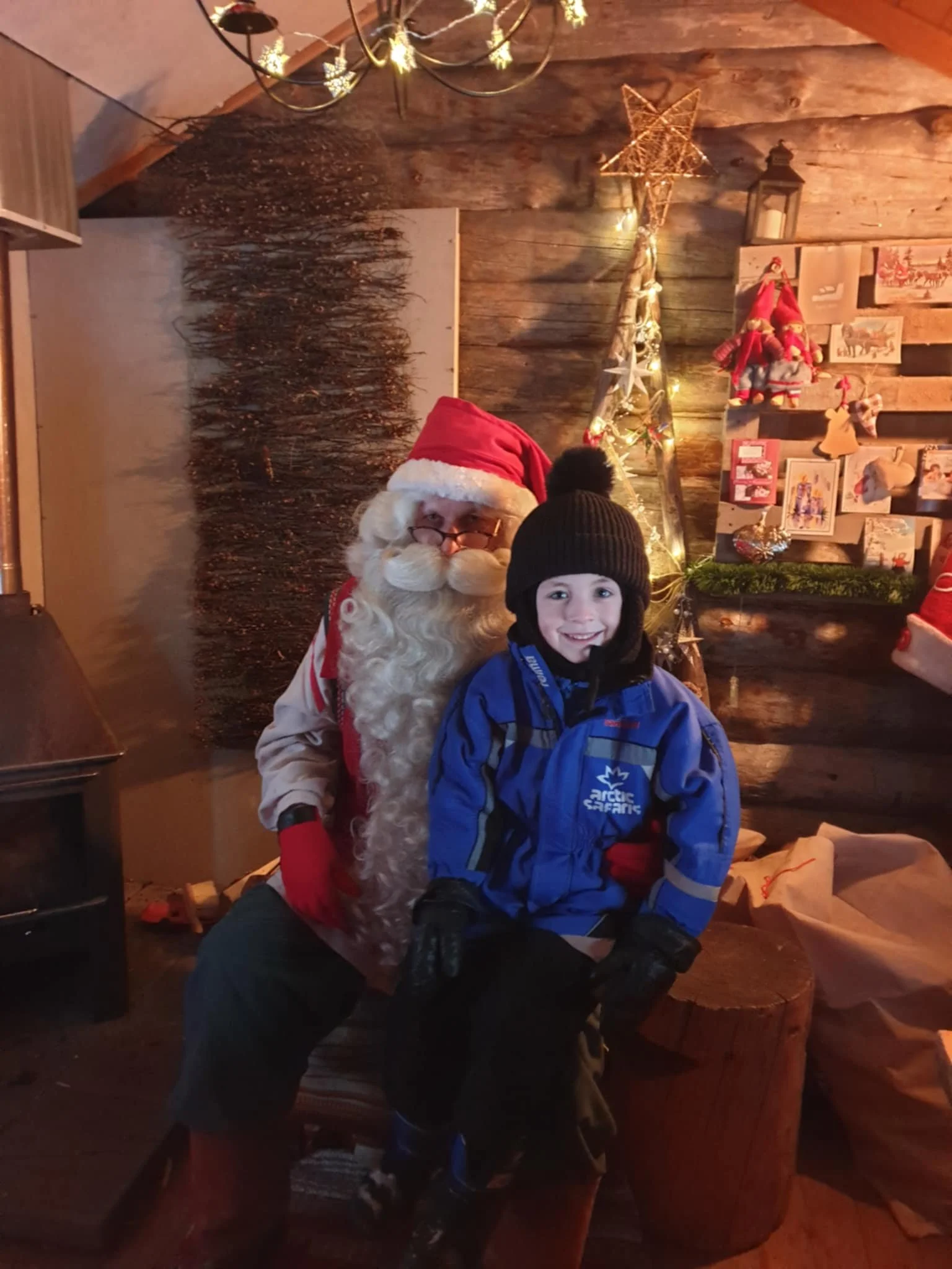 A boy sitting on Santa's lap in a cozy, wooden decorated room with Christmas ornaments and lights.