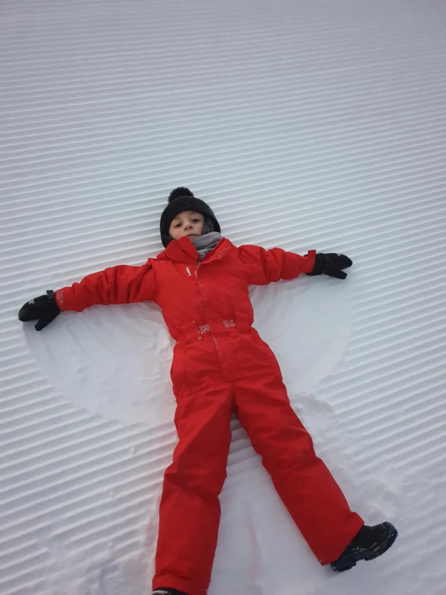 Child dressed in red snow suit, black hat, and gloves making a snow angel in snow with horizontal lines.