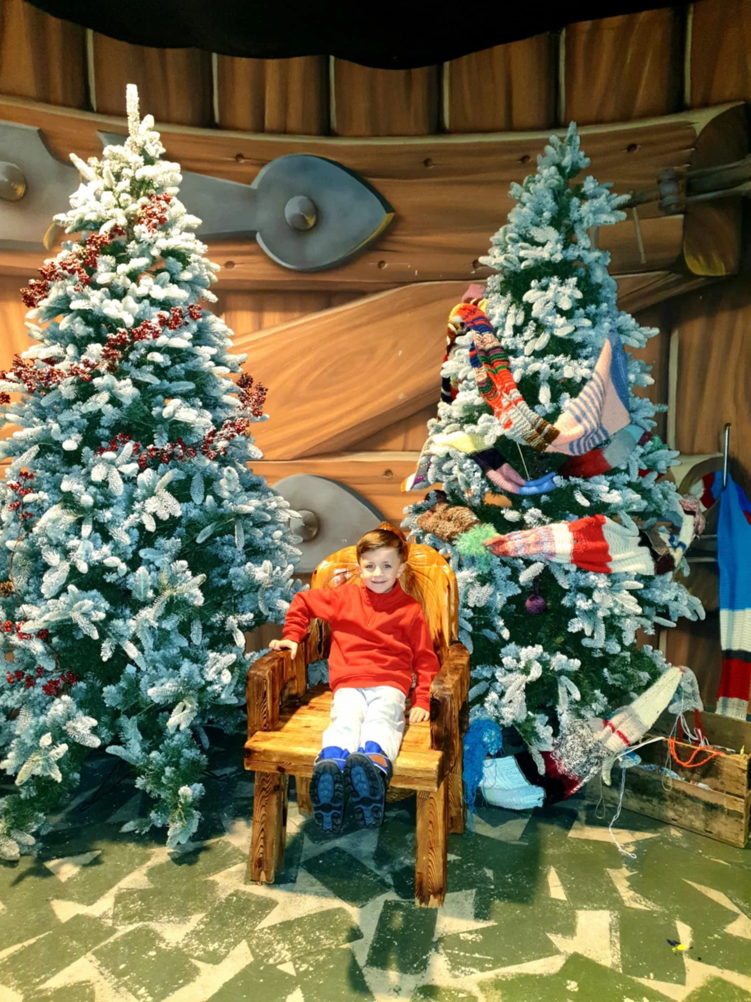 Young boy in red sweater sitting on a wooden chair between two decorated snow-covered Christmas trees with scarves hanging on one tree, wooden wall with nautical decor in the background, and snow-like decorations on the floor.