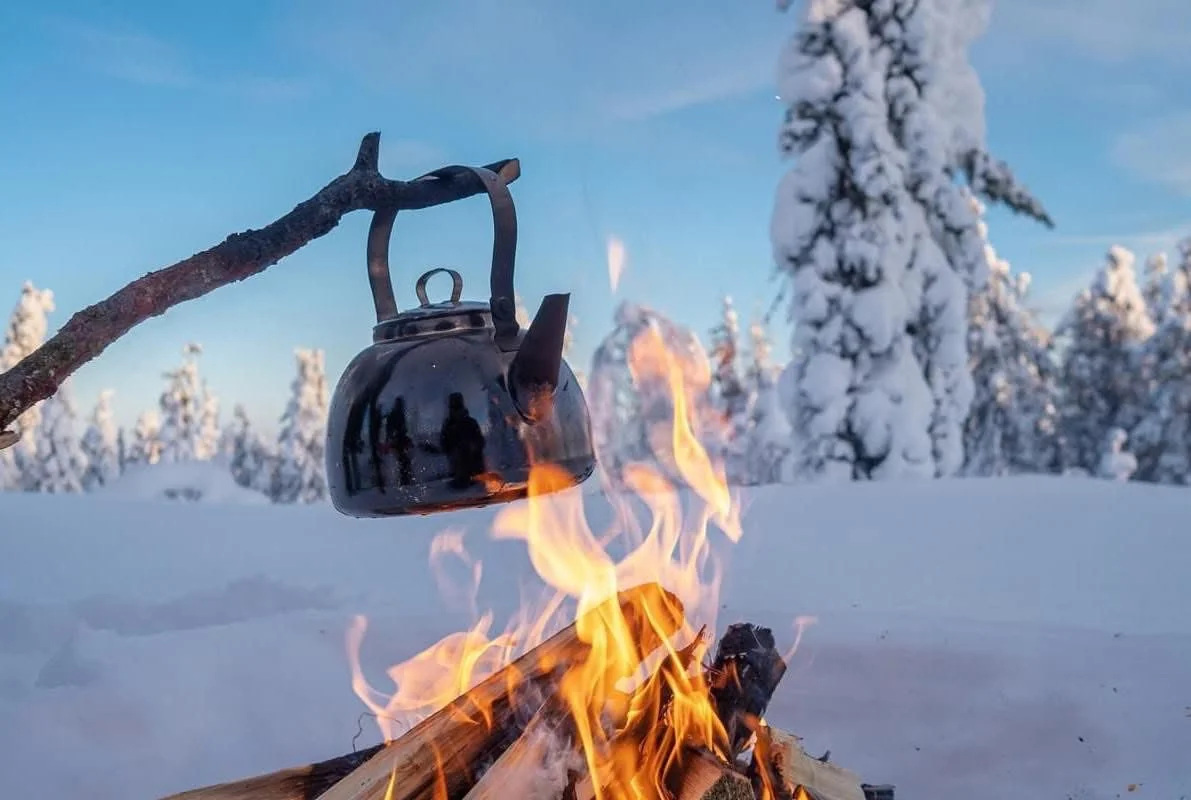 A black kettle hanging from a tree branch over an open campfire in a snowy landscape with snow-covered trees and a clear blue sky.