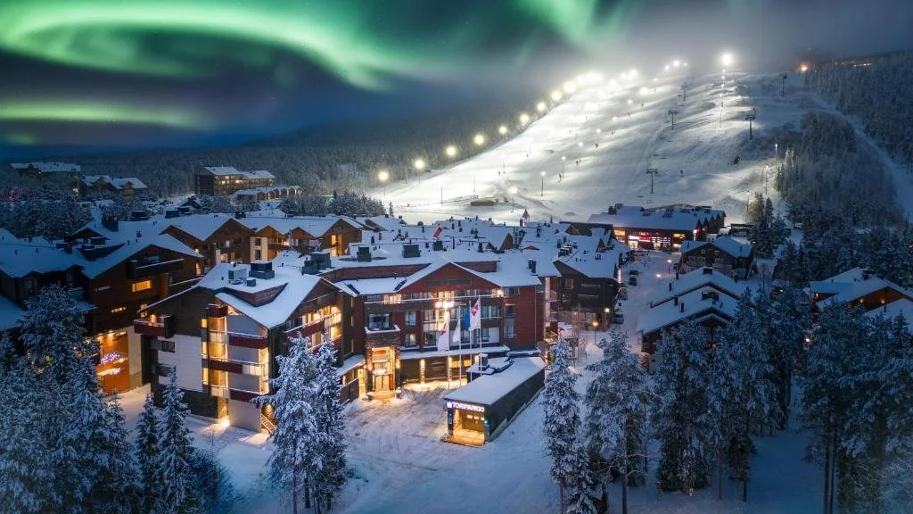 Snow-covered village at night with illuminated buildings, ski slopes, and northern lights in the sky.