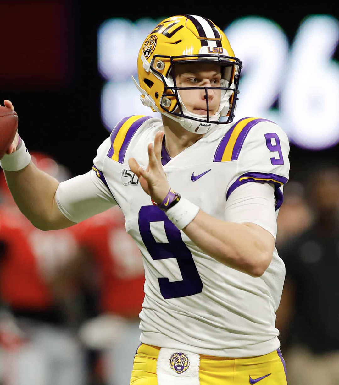 A young football player in a white and purple LSU uniform, wearing a yellow helmet with a tiger logo, is preparing to throw a football during a game.