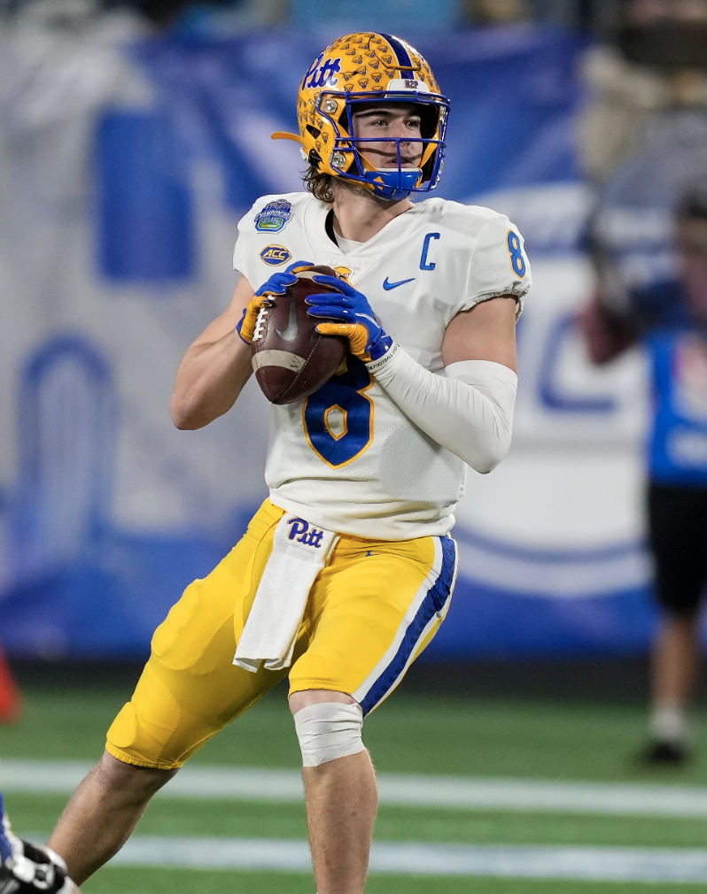 A college football player in a white and yellow uniform and blue helmet holding a football, standing on the field during a game.