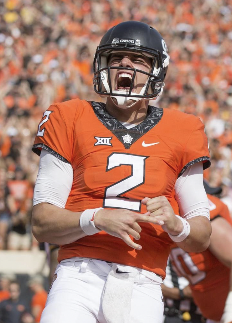 A football player celebrating on the field, wearing an orange jersey with the number 2, black helmet, and white pants, with a crowd in the background.