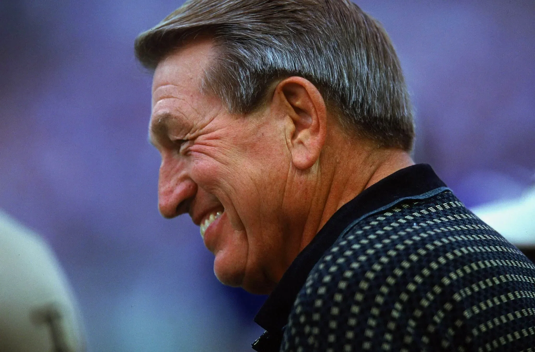 A smiling middle-aged man with brown hair and a dimpled smile, wearing a checkered patterned garment, captured in profile against a blurred background.