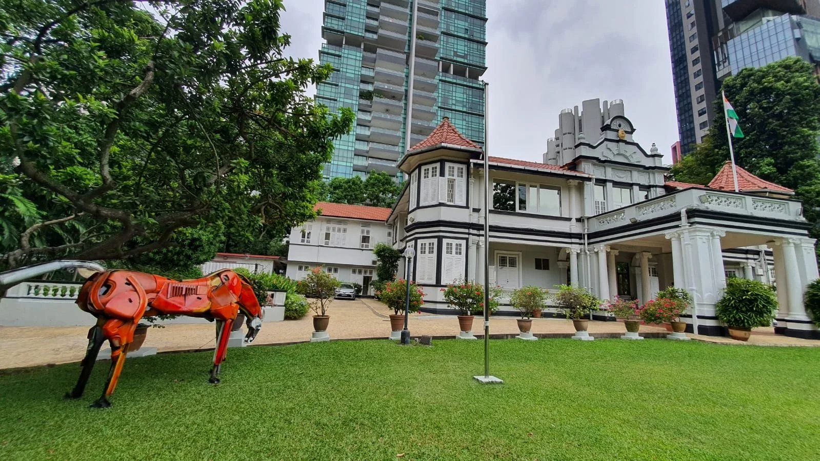 A white historic house with black accents and a red-tiled roof, surrounded by potted plants and lush greenery, in front of modern high-rise buildings. A flagpole with an Indian flag is also visible.