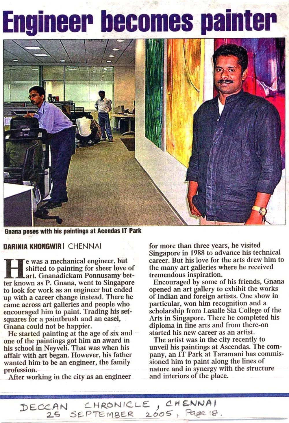 A man stands in an art gallery with colorful paintings on the wall, smiling and holding a small object, with office workers and art pieces visible in the background.