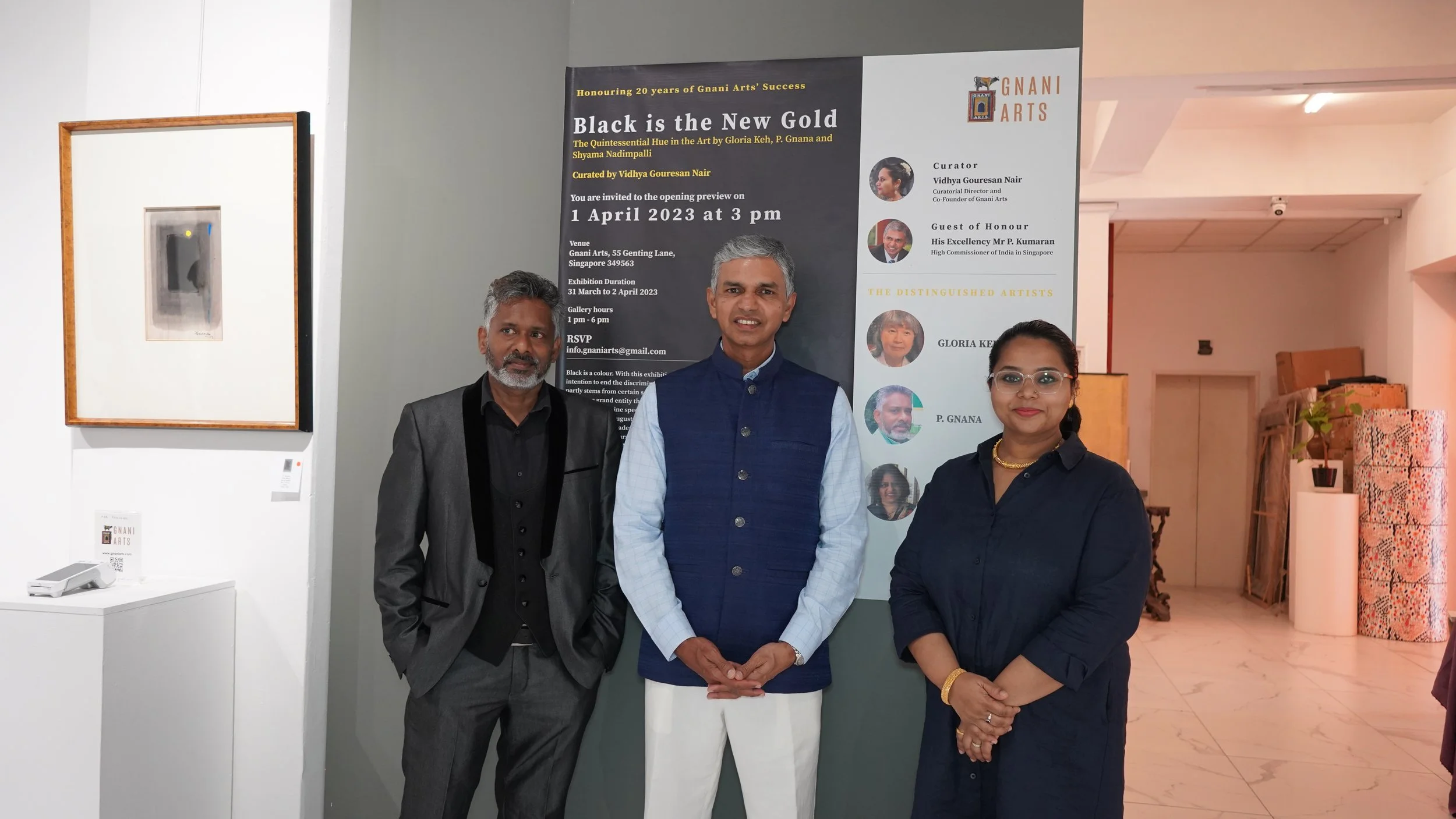Three people standing indoors in front of a poster about an art exhibition titled "Black is the New Gold" at Gnani Arts, celebrating 20 years of the gallery's success, with artwork displayed on the wall to the side.