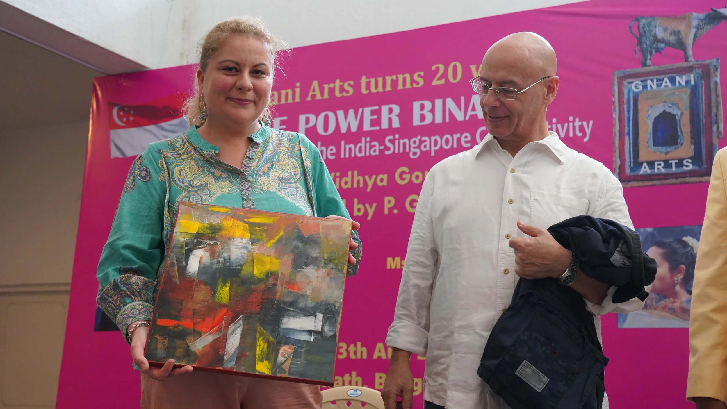 A woman in a colorful traditional dress holding an abstract painting stands next to a man in a white shirt at an art event, with a pink banner in the background celebrating 20 years of Gnani Arts.