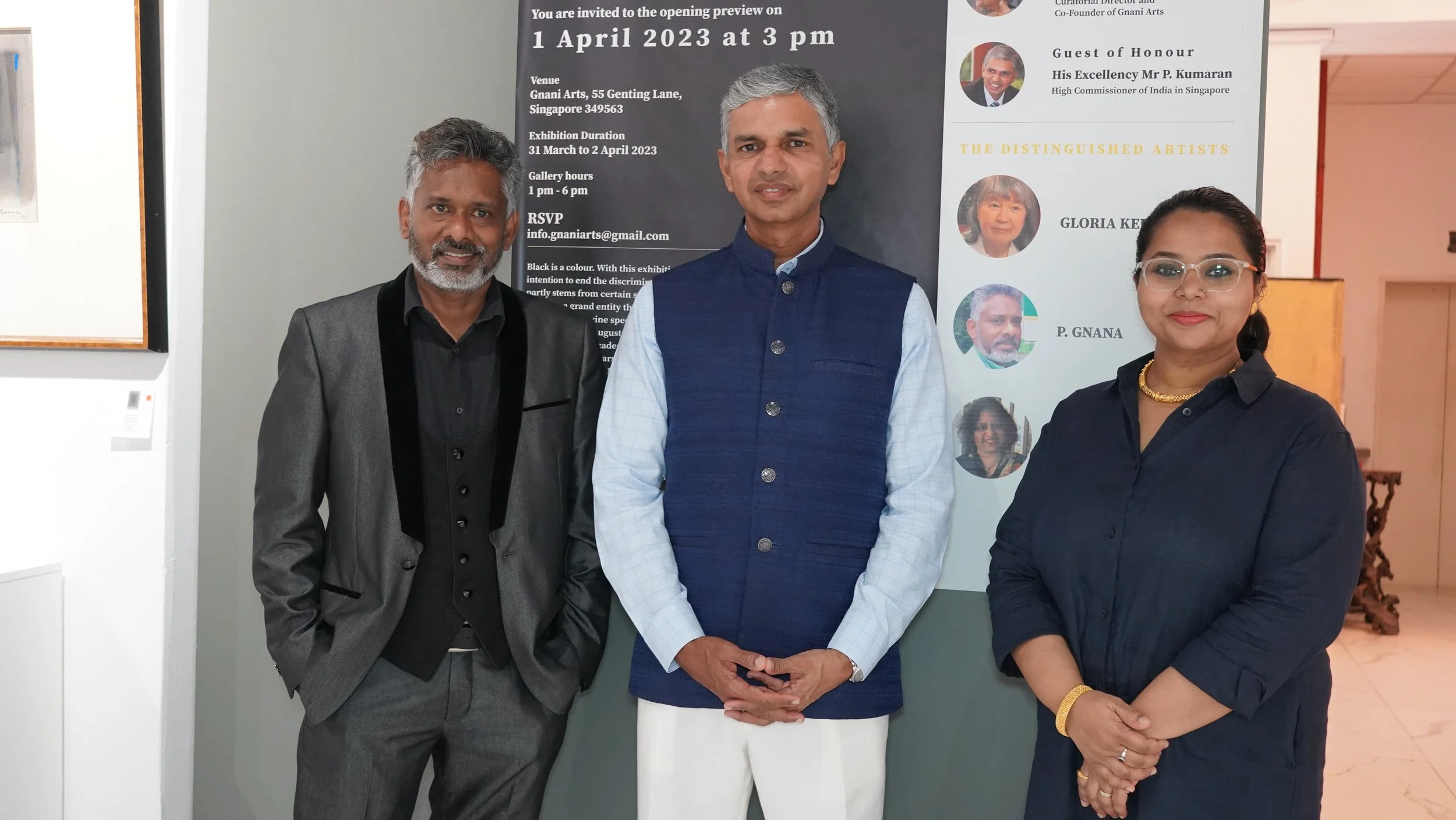 Three people standing in front of an event poster at an art exhibition. The man on the left has gray hair and a beard, and is wearing a gray suit with black accents. The man in the middle has short gray hair, is dressed in a blue vest over a light bl