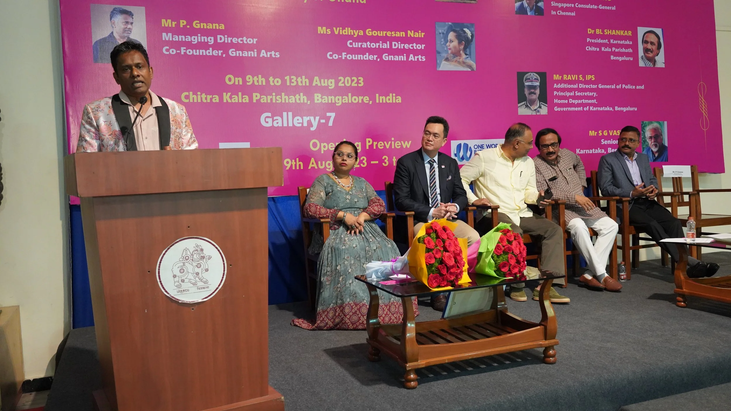 A man in a floral blazer is speaking at a podium with a pink backdrop behind him. The backdrop displays event details and photographs of several individuals, indicating a formal event or conference. Six people are seated to the right of the speaker, 